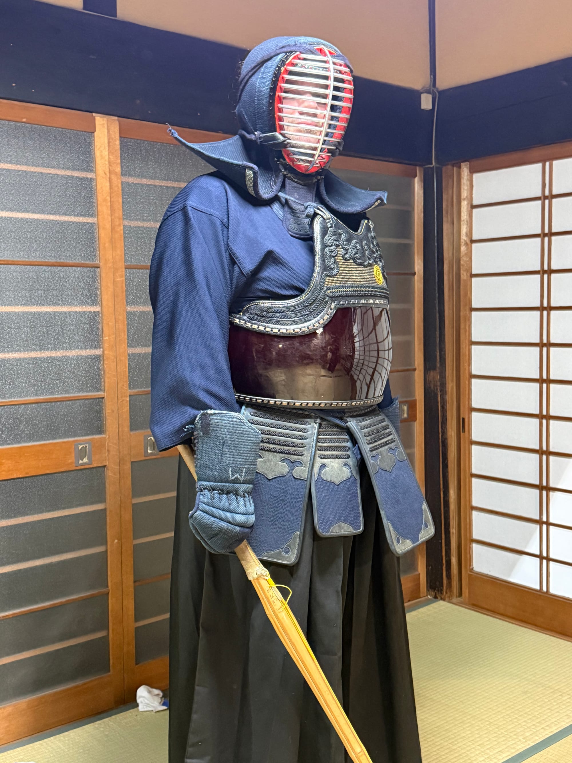 Visitor wearing full kendo armour in a traditional tatami room in Kyoto, holding a bamboo shinai, with shoji screens and martial arts equipment visible in the background.