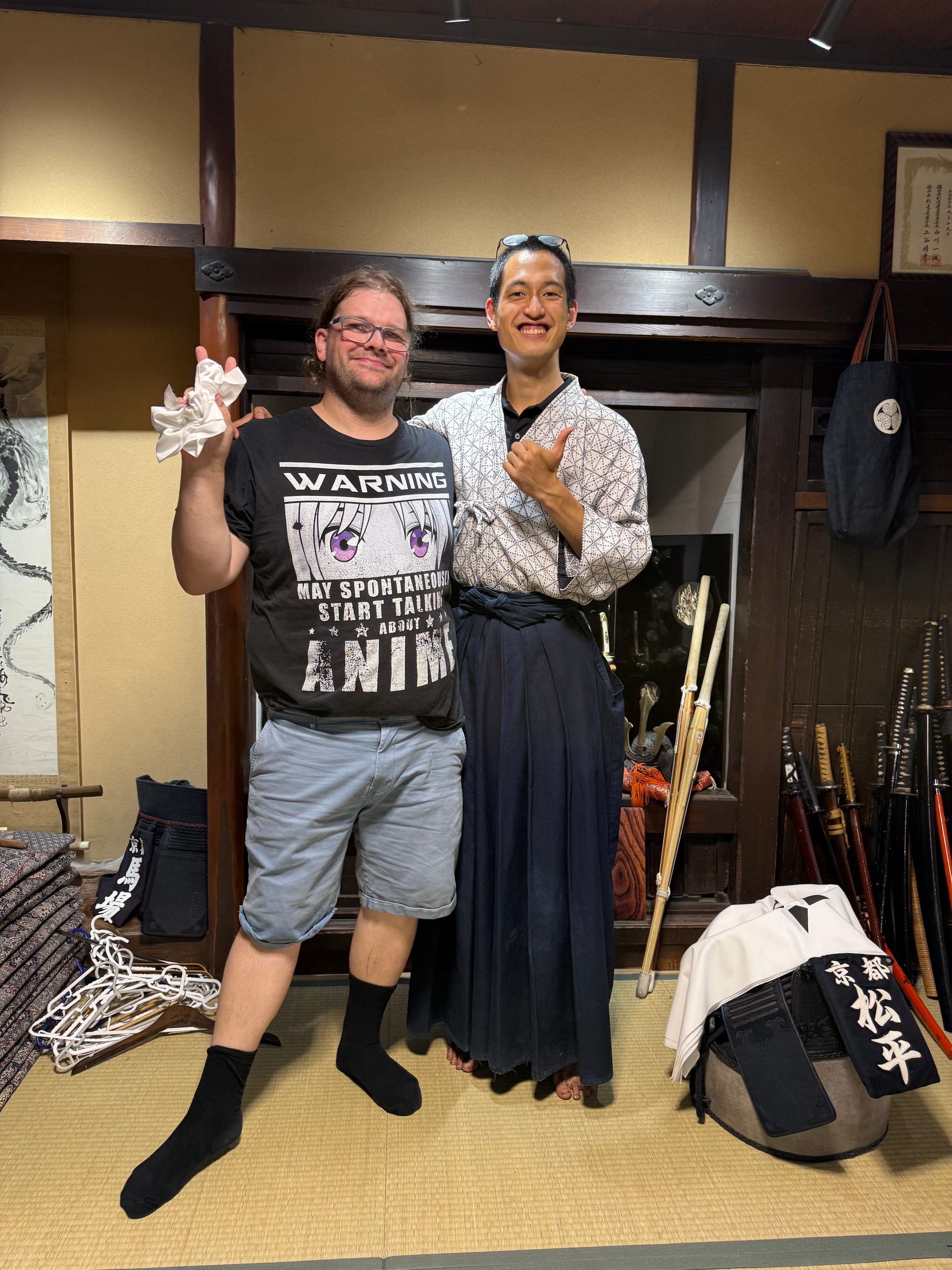 Two visitors pose inside a traditional Japanese room, with one wearing hakama-style clothing, surrounded by kendo equipment, swords and martial arts displays in Kyoto.