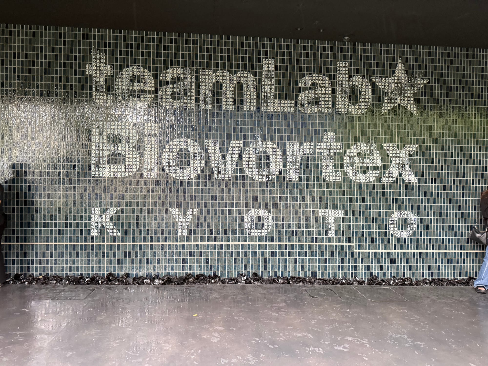 TeamLab Biovortex Kyoto entrance wall featuring cascading water over blue mosaic tiles, with the venue name displayed in large white lettering above a reflective floor.