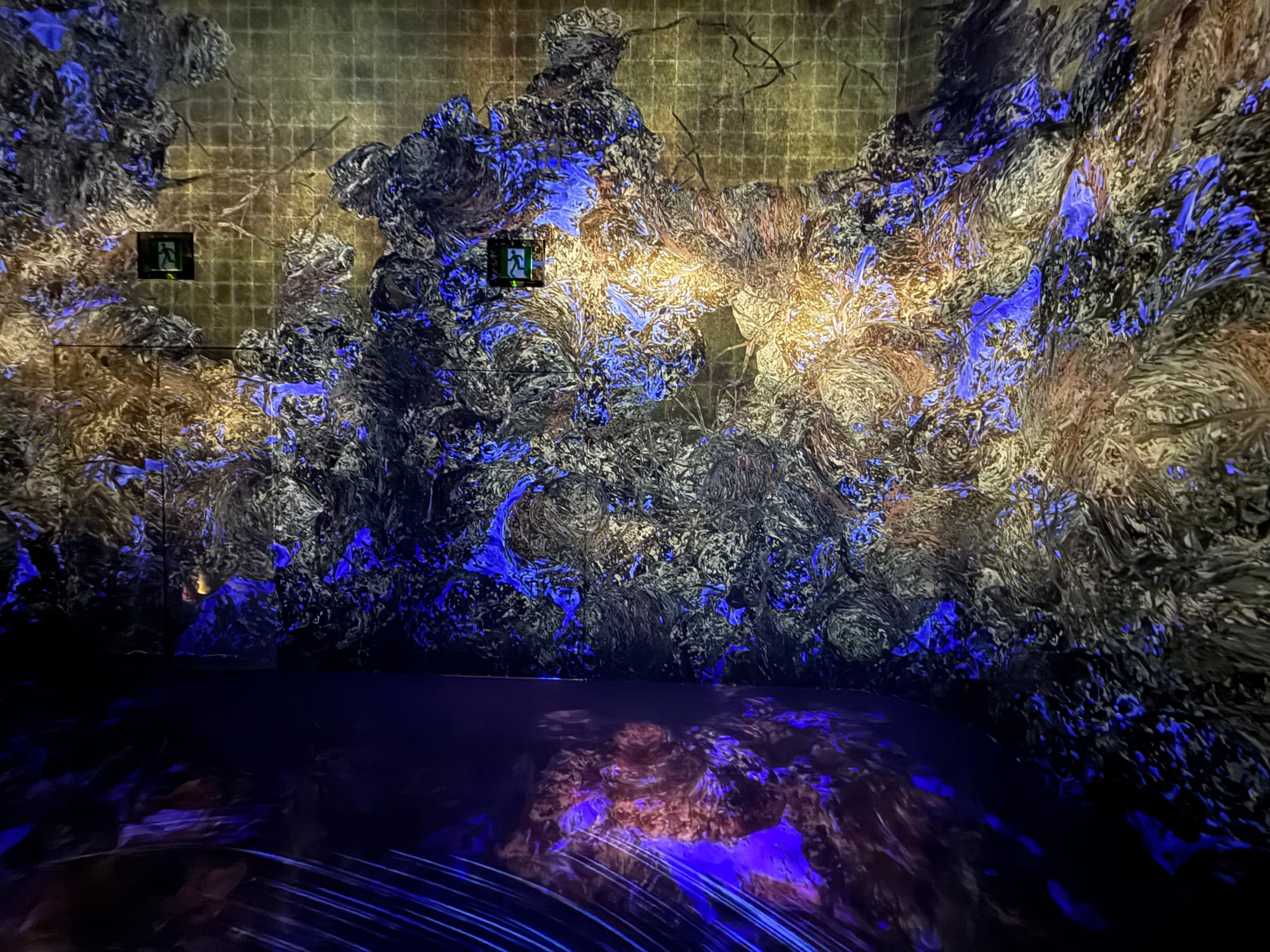 Immersive digital artwork at teamLab Biovortex Kyoto, featuring swirling blue, gold and black abstract forms projected across the walls and reflected on the glossy floor.