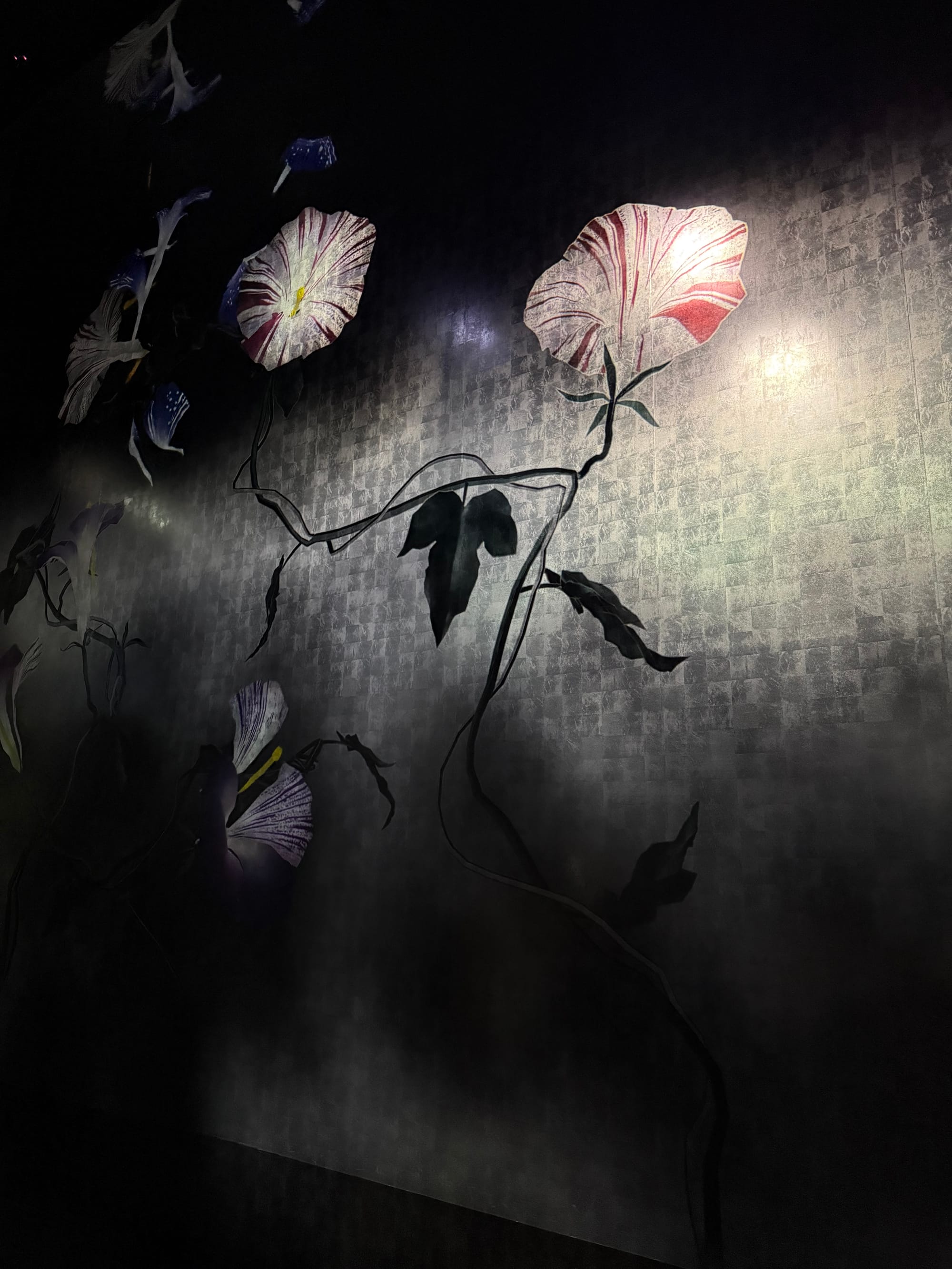 Large illuminated flower installation at teamLab Biovortex Kyoto, featuring oversized morning glory blossoms and twisting vines suspended against a dark textured wall.