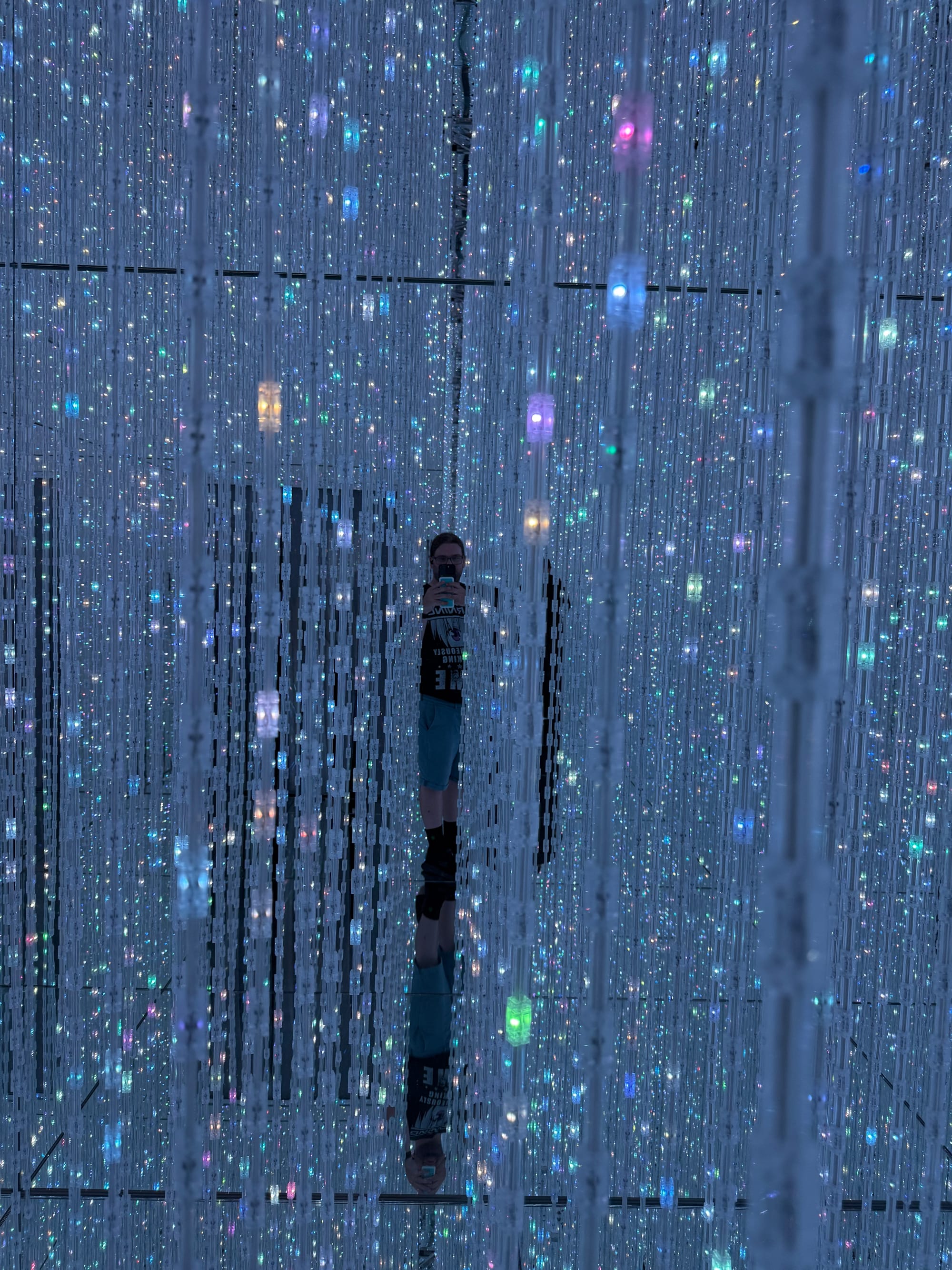 Visitor standing within an immersive light installation at teamLab Biovortex Kyoto, surrounded by thousands of suspended illuminated strands and mirrored reflections in every direction.