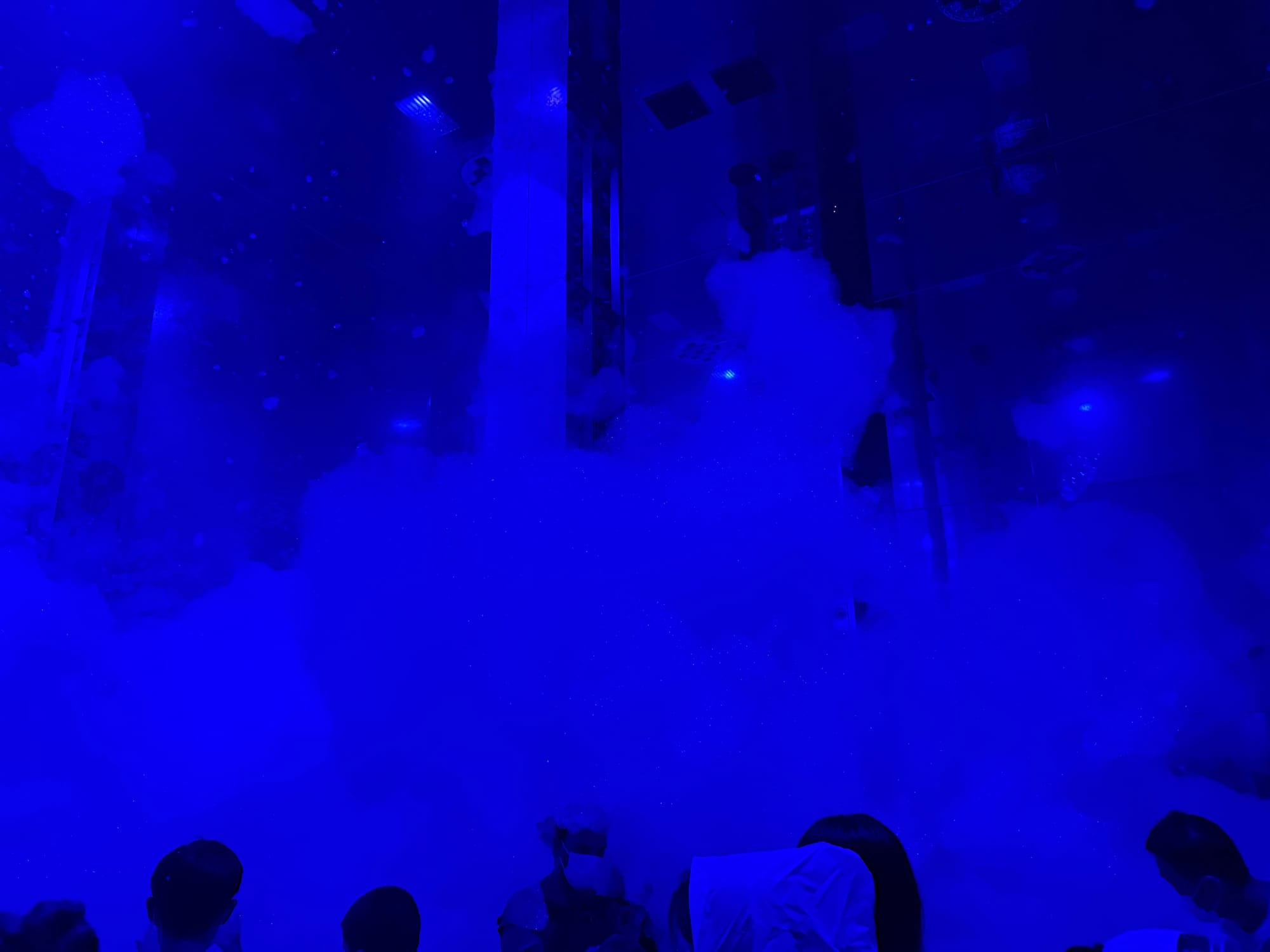 Massless Amorphous Sculpture at teamLab Biovortex Kyoto, with visitors gathered around drifting cloud-like forms illuminated in deep blue light and reflected by mirrored surfaces.