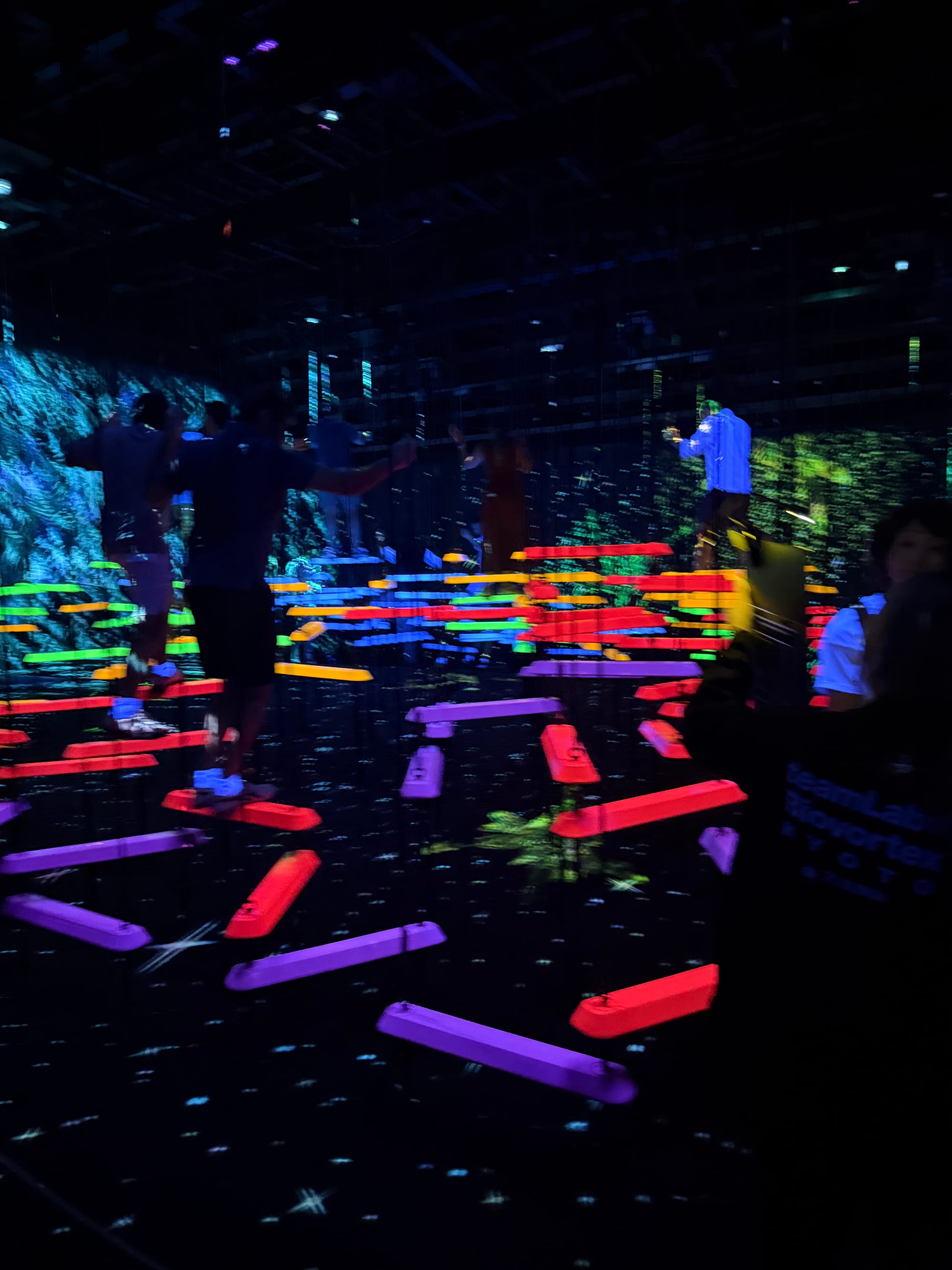 Visitors cross illuminated balancing beams in an interactive teamLab installation, with colourful glowing platforms, mirrored walls and animated digital projections.