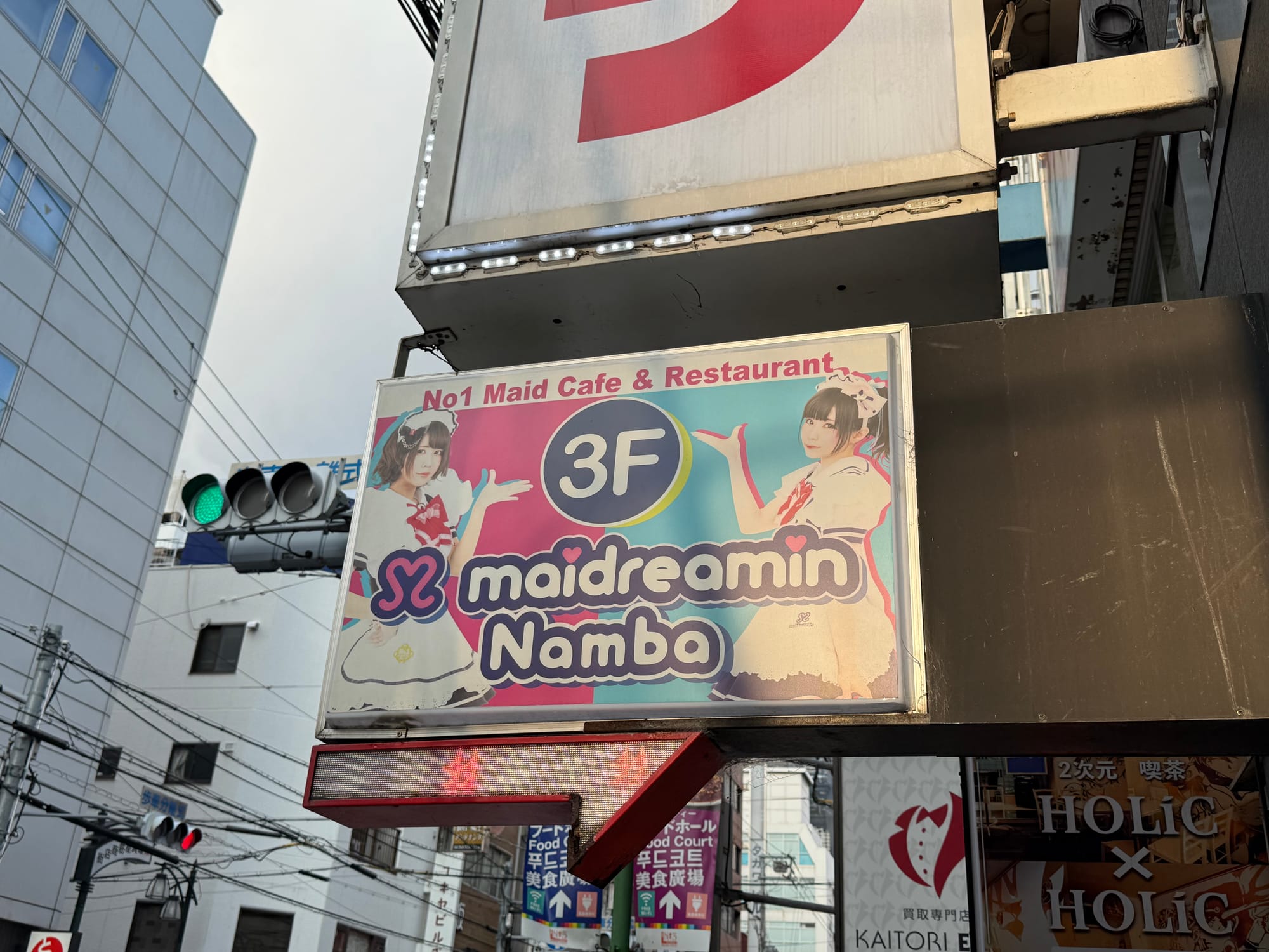 Sign for maidreamin Namba in Osaka's Namba district, advertising the maid café on the third floor, with colourful branding, street signs and surrounding city buildings.