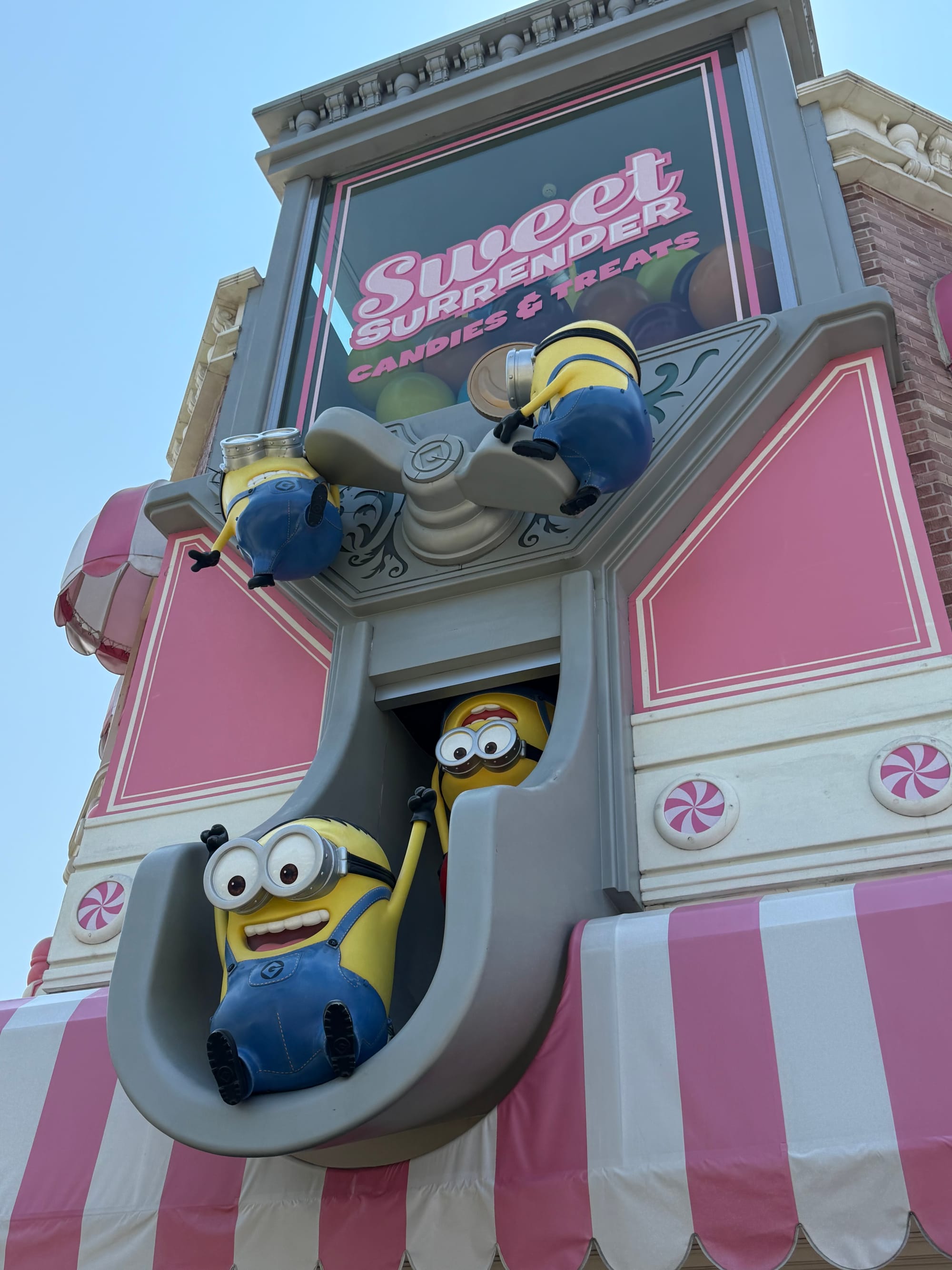 Minion figures decorating the Sweet Surrender candy shop at Universal Studios Japan, with characters sliding from a giant scoop above the pink-and-white confectionery-themed façade.
