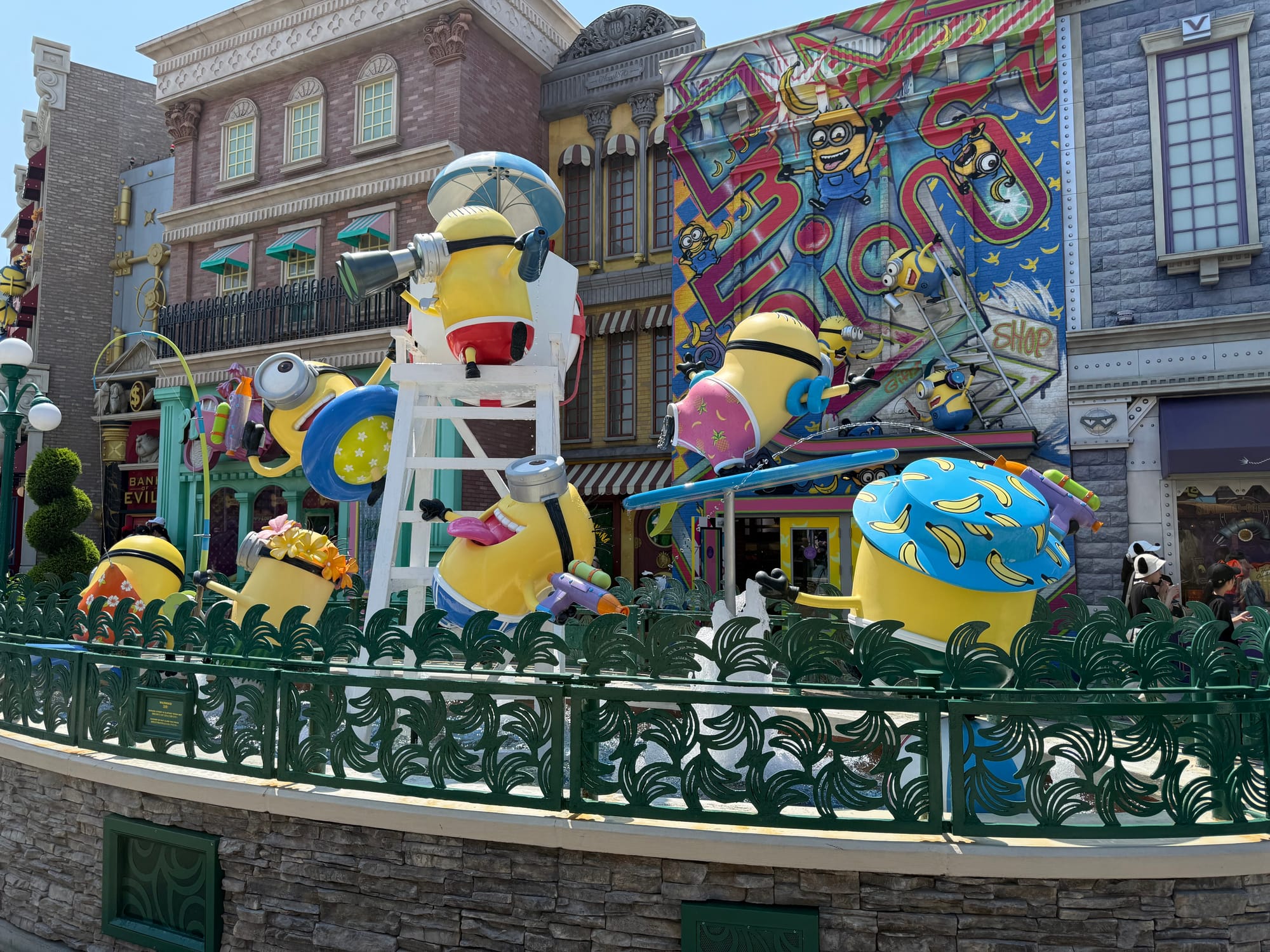 Minion-themed fountain at Universal Studios Japan, featuring multiple Minion figures in colourful summer outfits playfully interacting around water jets, props and a graffiti-covered backdrop.