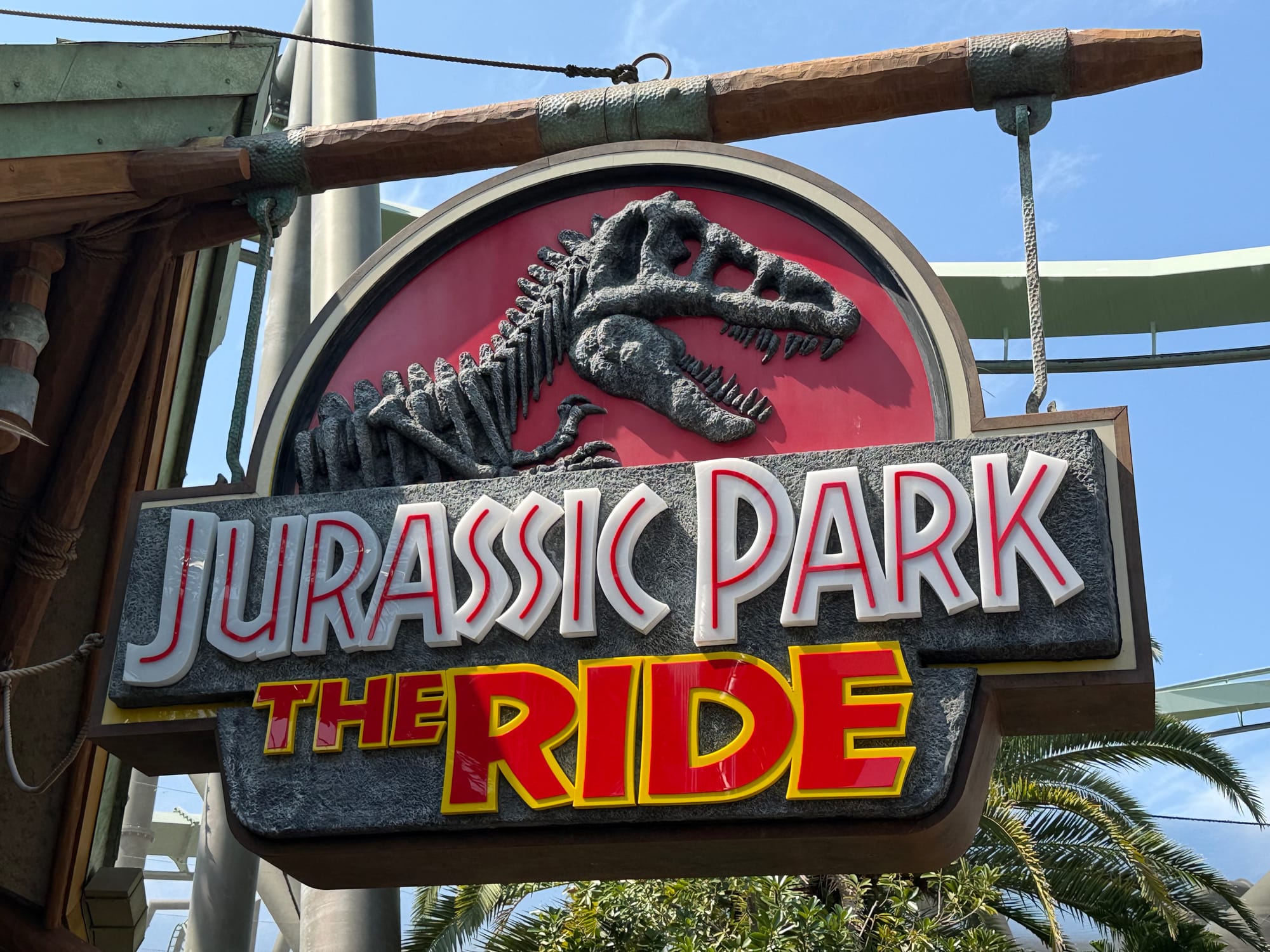 Sign for Jurassic Park: The Ride at Universal Studios Japan, featuring the iconic Tyrannosaurus rex skeleton logo mounted above the attraction name with coaster track visible behind.