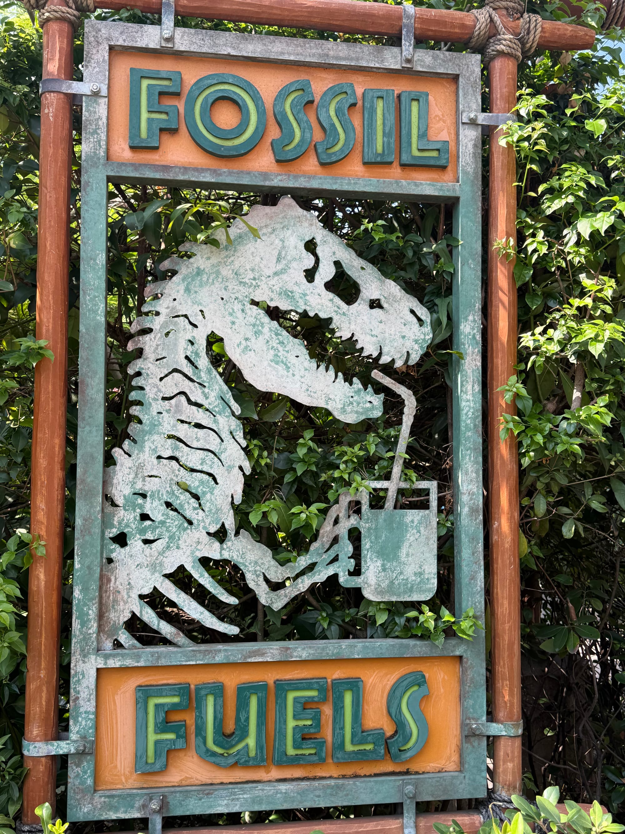 Fossil Fuels sign in Jurassic Park at Universal Studios Japan, featuring a metal Tyrannosaurus rex skeleton drinking from a fuel can, surrounded by lush greenery.
