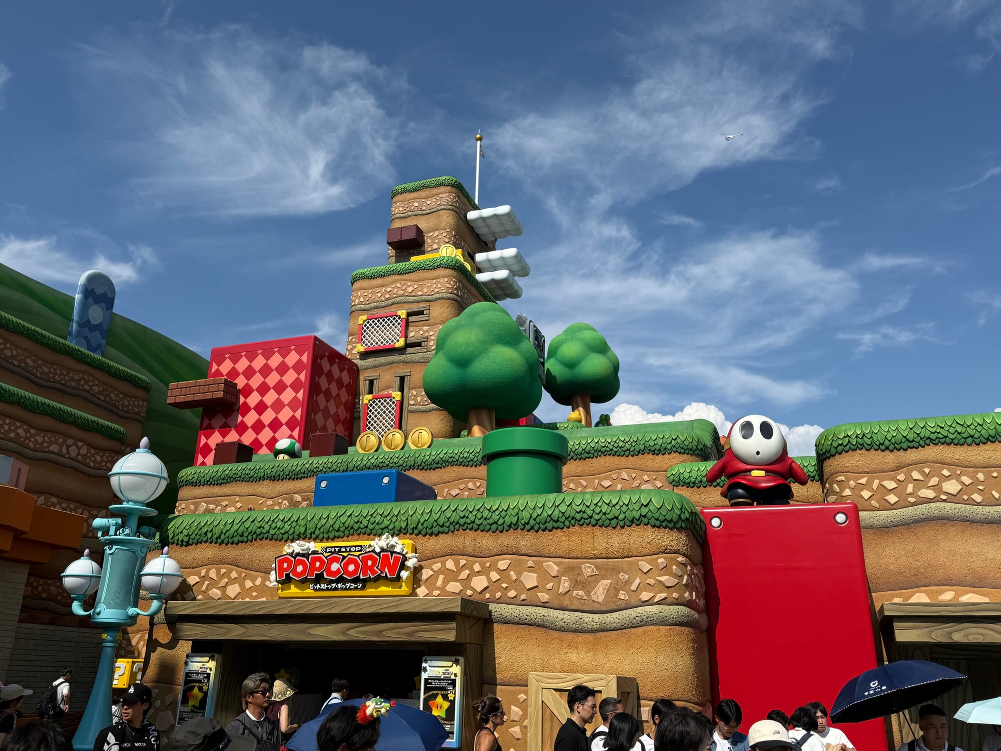 Super Nintendo World at Universal Studios Japan, featuring layered Mario-themed scenery, green warp pipes, floating coin blocks, a Shy Guy figure and crowds below.