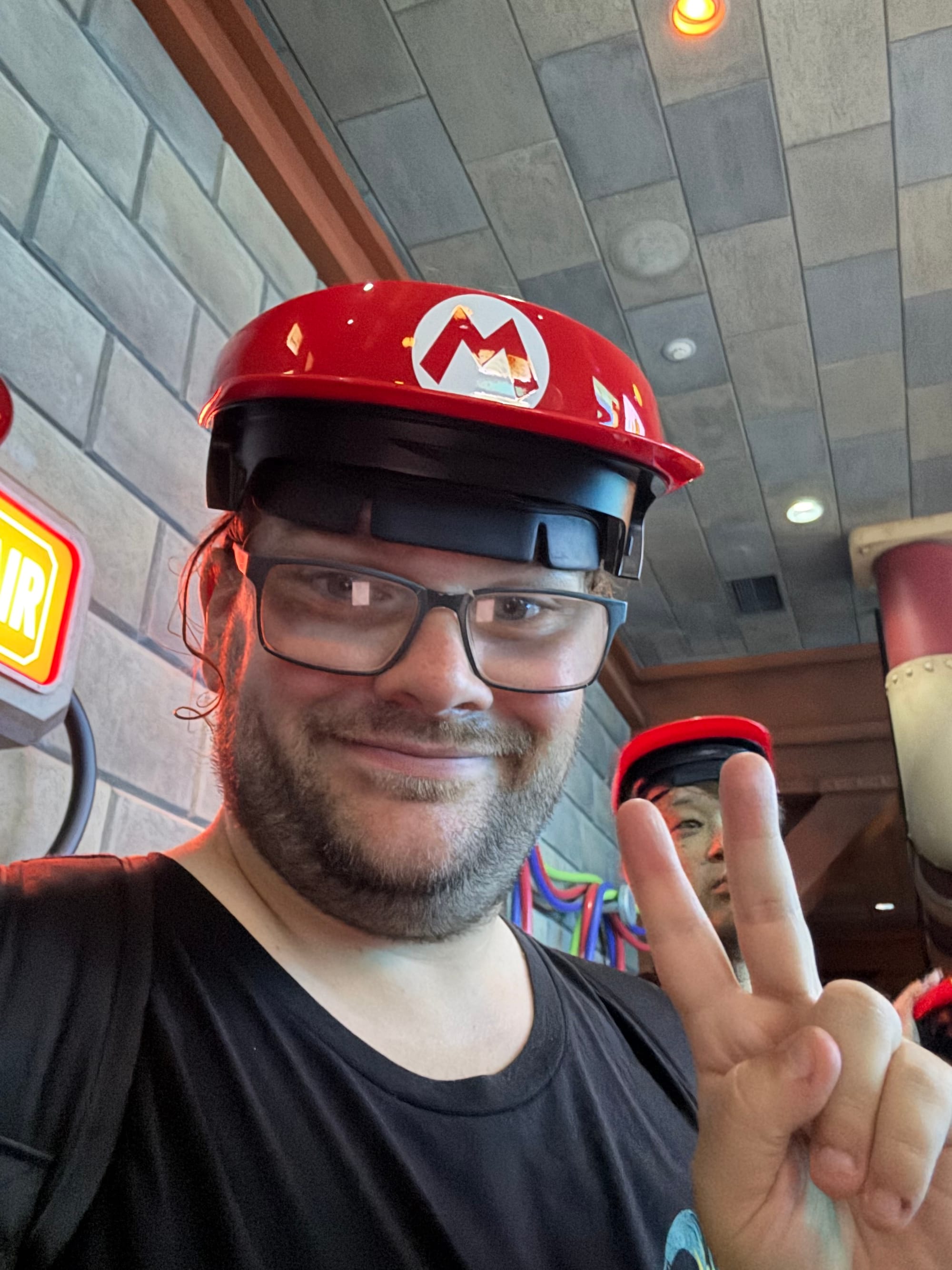 Visitor wearing a Mario Kart augmented reality visor and Mario-themed helmet inside Mario Kart: Koopa's Challenge at Super Nintendo World, posing with a peace sign before boarding.