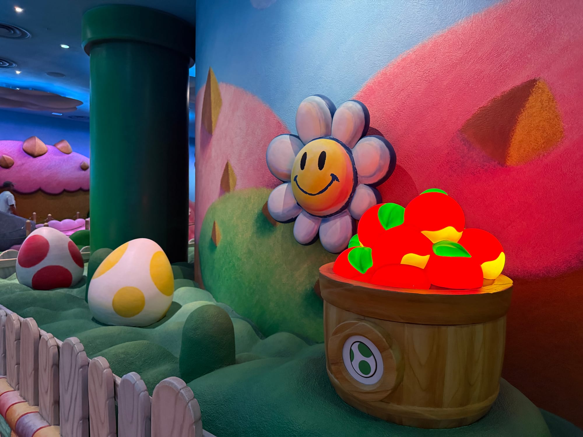 Colourful themed scenery in the queue for Yoshi's Adventure at Super Nintendo World, featuring Yoshi eggs, smiling flowers, oversized fruit and a green warp pipe.