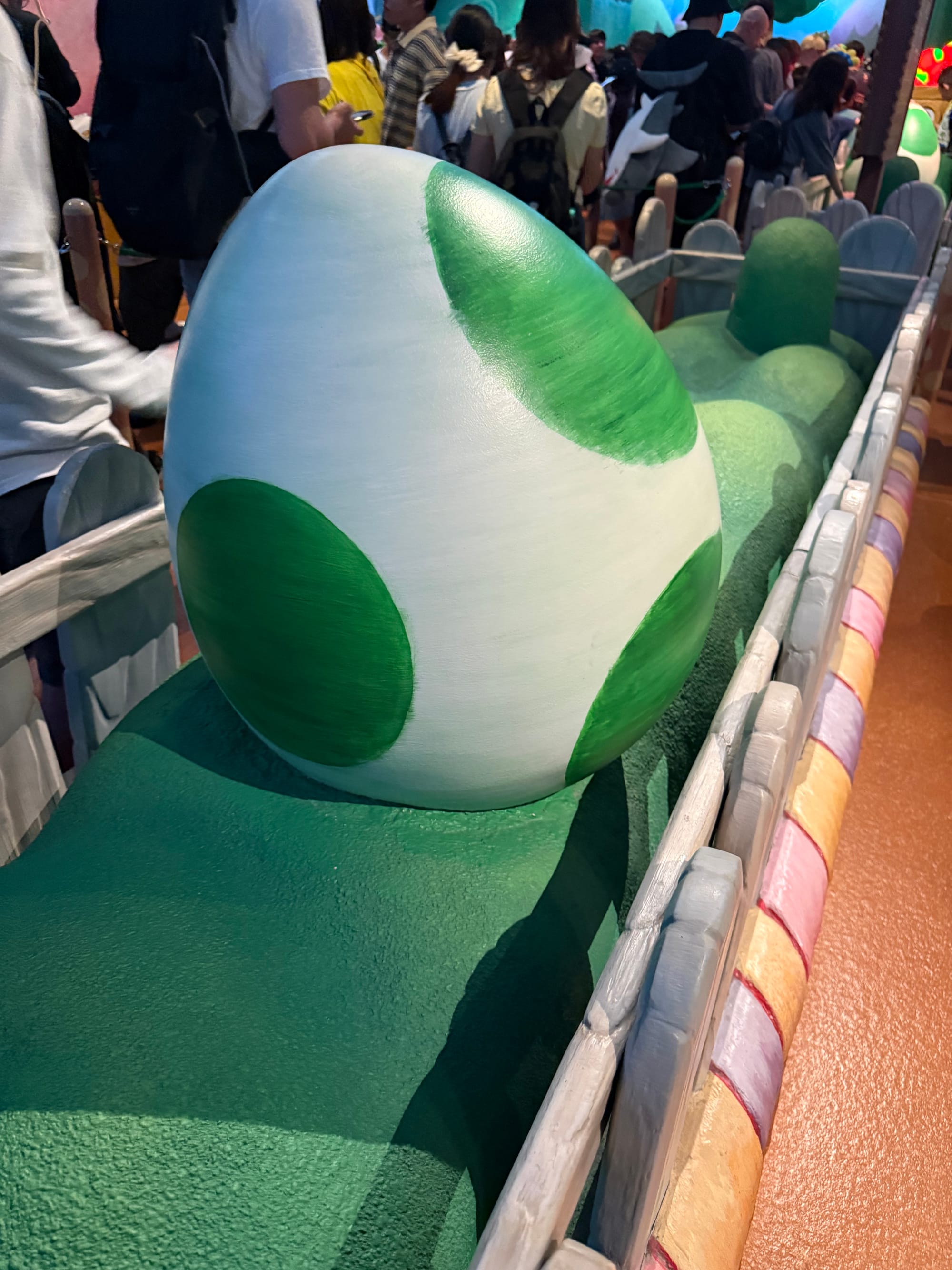 Large Yoshi egg with green spots displayed in the queue for Yoshi's Adventure at Super Nintendo World, surrounded by colourful themed scenery and crowds of waiting guests.