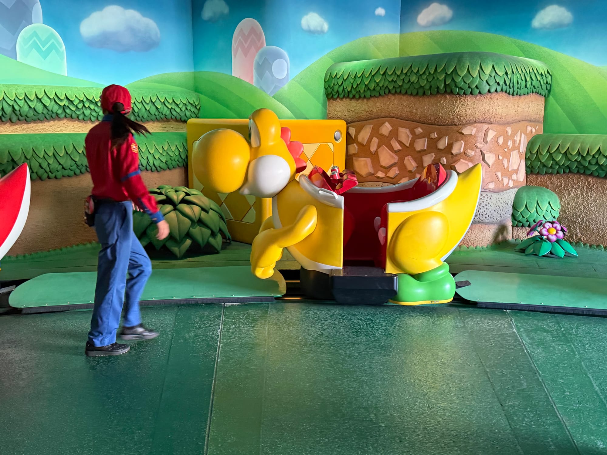 Yellow Yoshi ride vehicle at the loading platform for Yoshi's Adventure in Super Nintendo World, with colourful Mushroom Kingdom scenery and a team member preparing guests to board.
