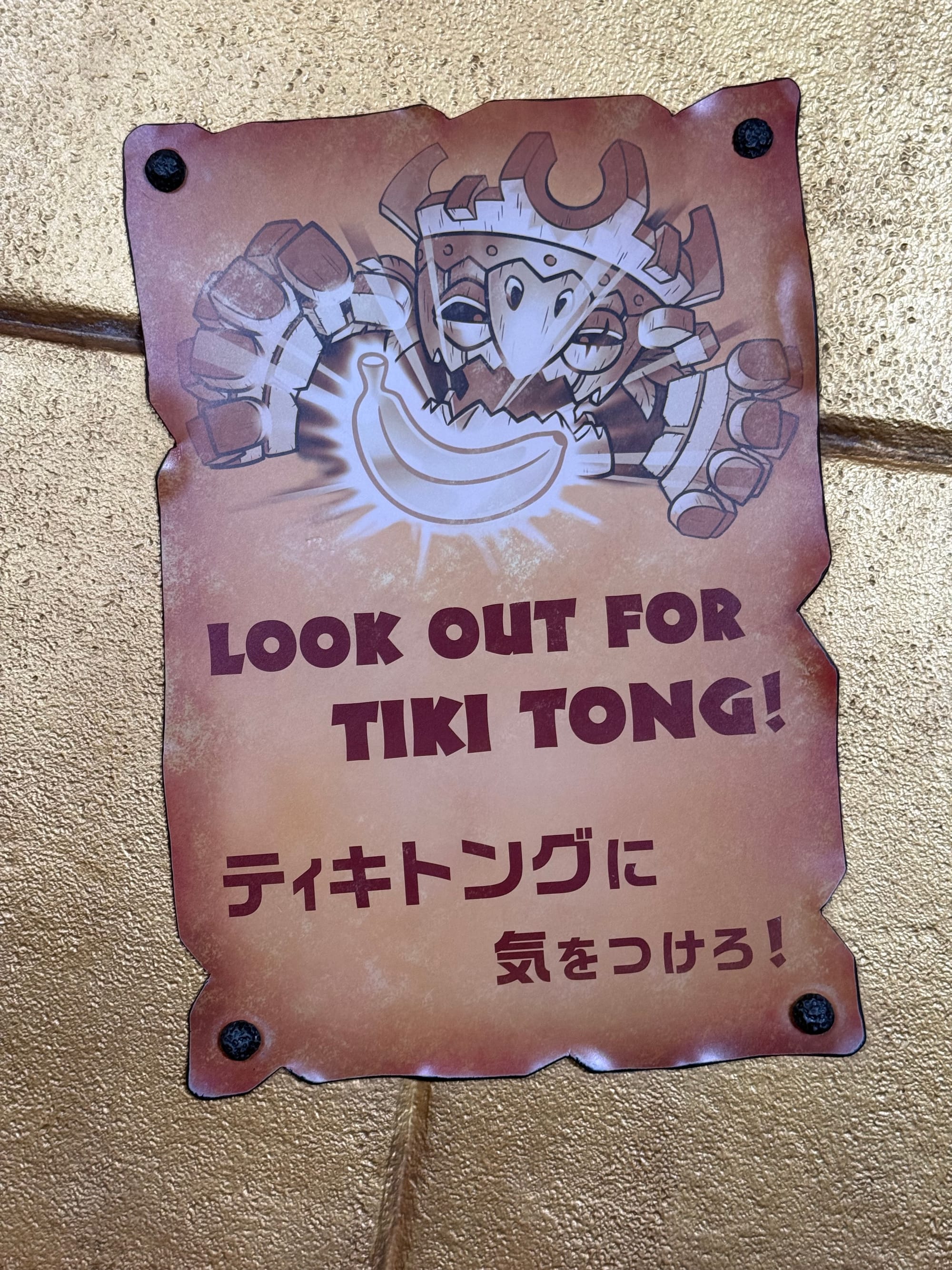 Warning sign in the Mine Cart Madness queue at Donkey Kong Country, depicting Tiki Tong reaching for a banana, with English and Japanese text alerting riders to the jungle villain.