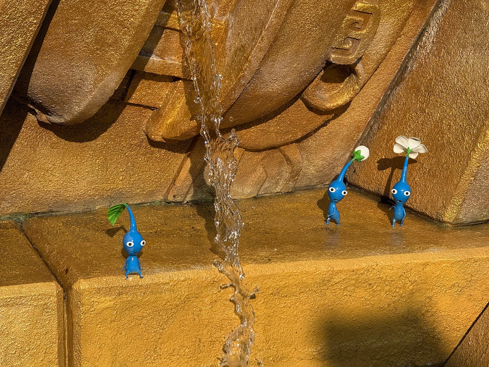 Blue Pikmin figures beside a small waterfall in Donkey Kong Country at Universal Studios Japan, standing among golden temple ruins with leaf and flower sprouts growing from their heads.