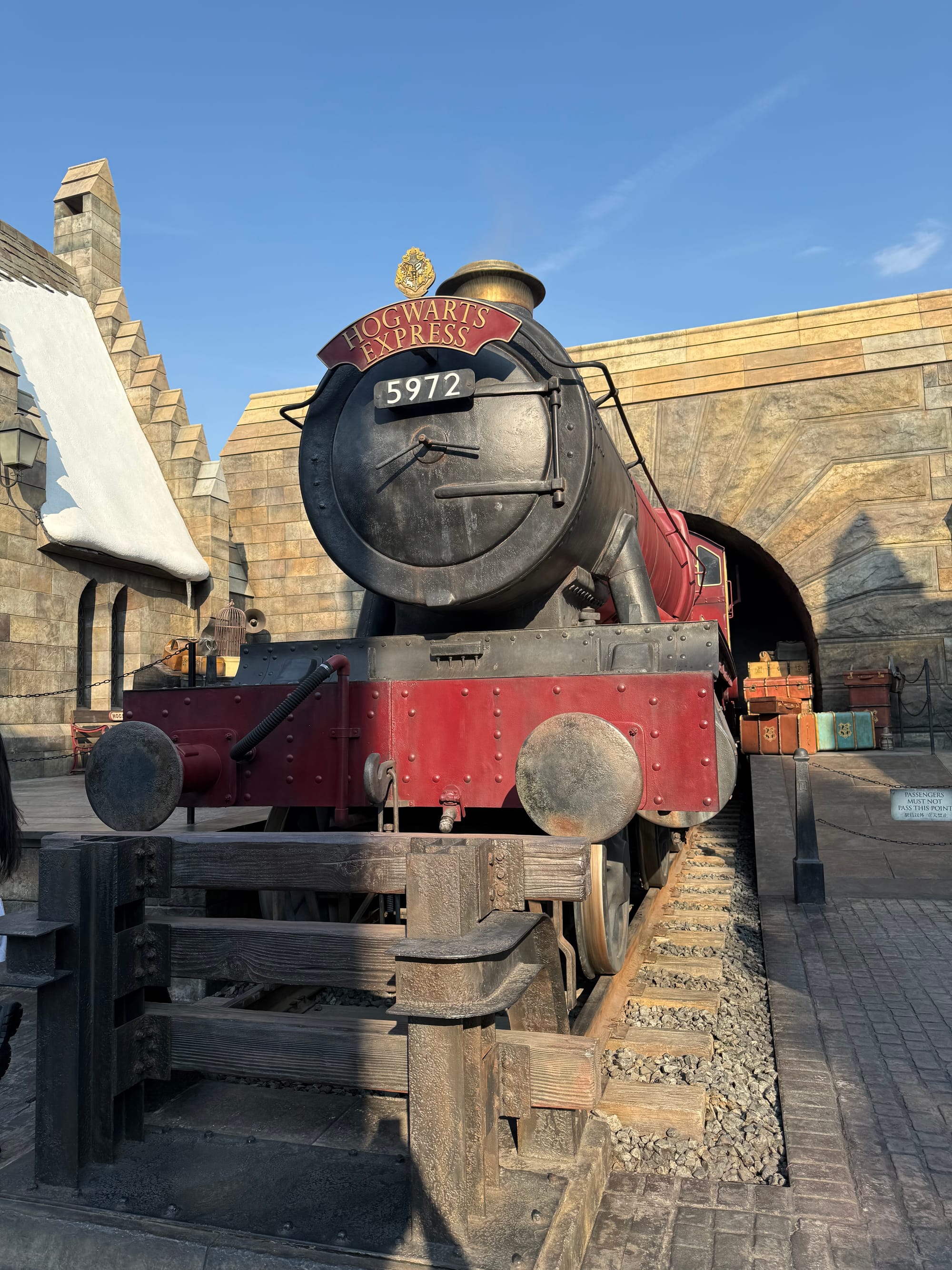 Hogwarts Express steam locomotive at The Wizarding World of Harry Potter in Universal Studios Japan, emerging from a tunnel beside Hogsmeade Station beneath a clear blue sky.