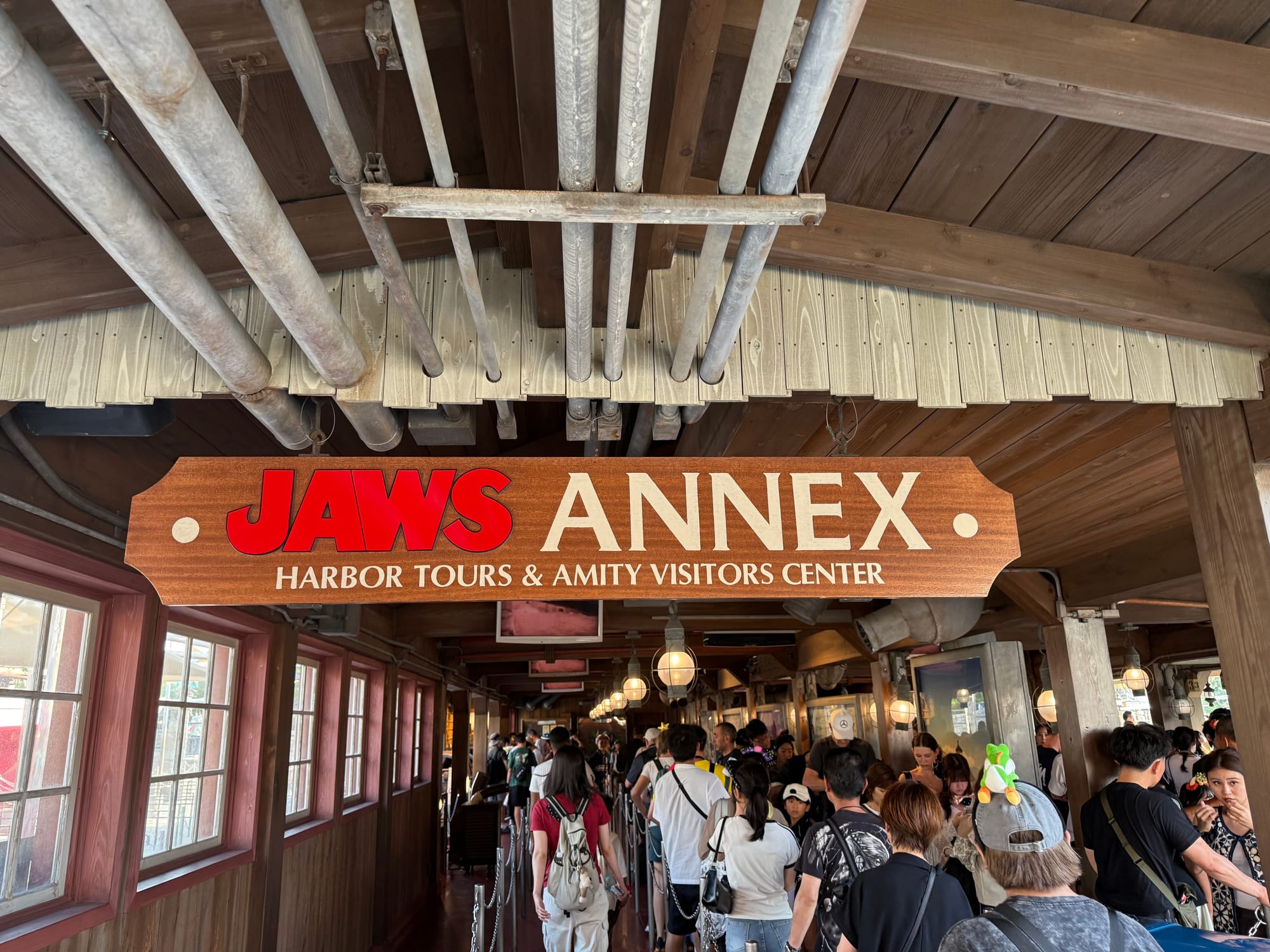 Queue entrance for Jaws at Universal Studios Japan, featuring a wooden sign reading "JAWS ANNEX: Harbor Tours & Amity Visitors Center" above a crowded themed walkway.