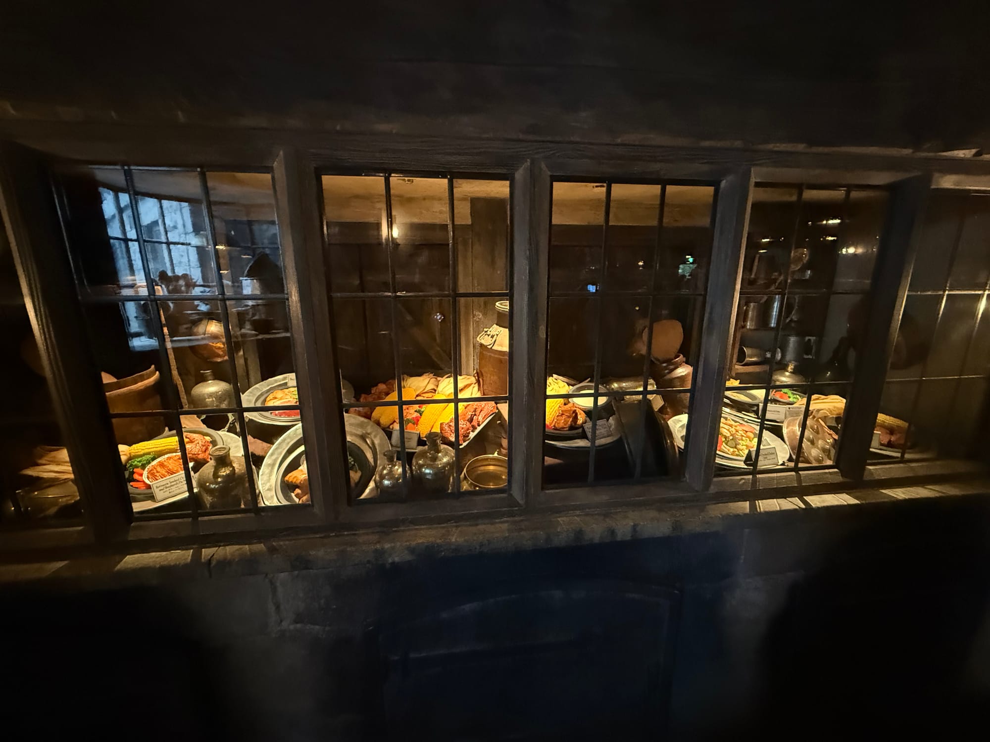 Display of food and drink props seen through a leaded window at the Three Broomsticks in The Wizarding World of Harry Potter, featuring platters of roast meals, bread and tankards.