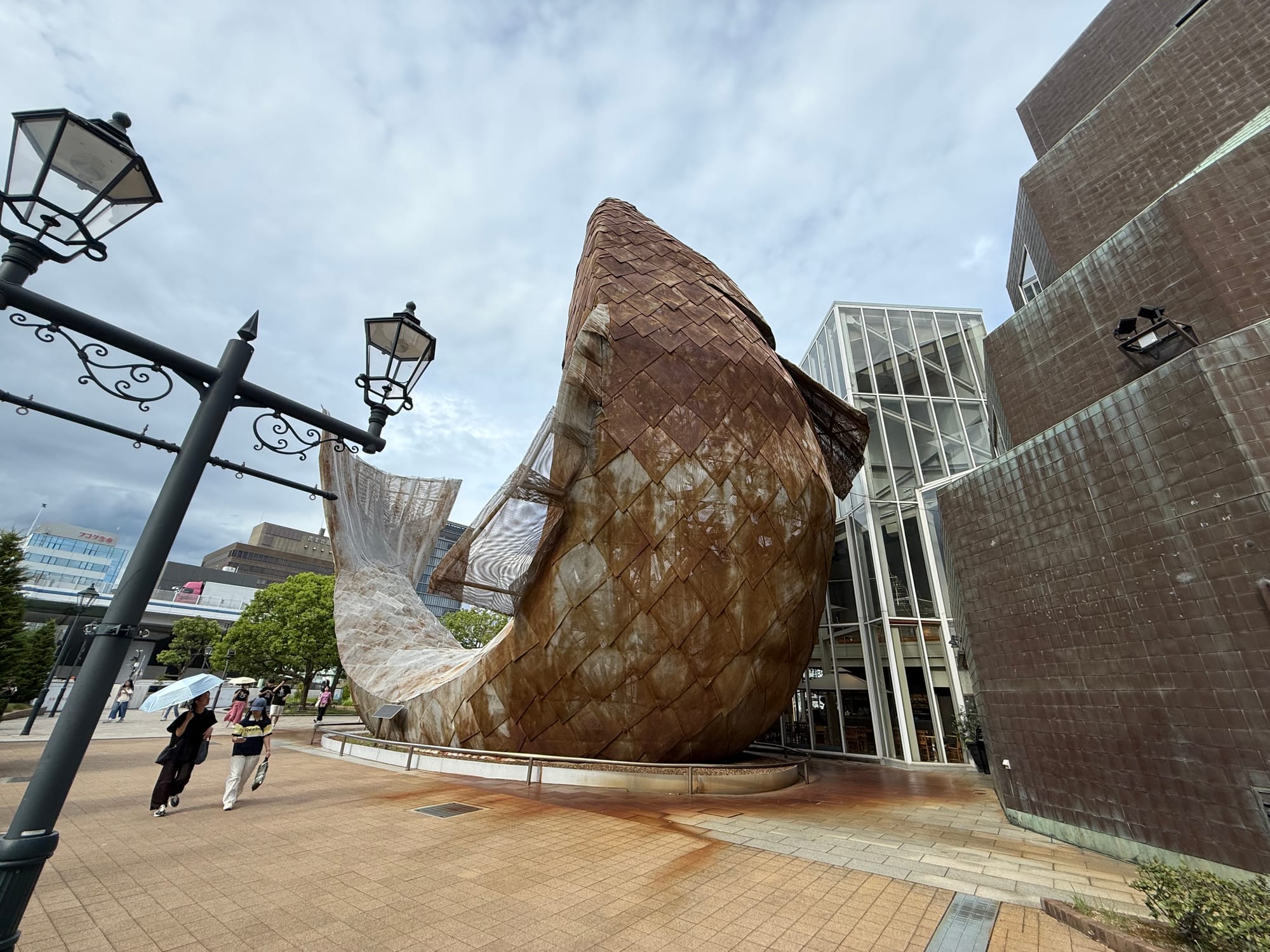 Fish Dance sculpture at Kobe Harborland, featuring a giant fish-shaped structure clad in copper-coloured scales beside the waterfront under an overcast sky.