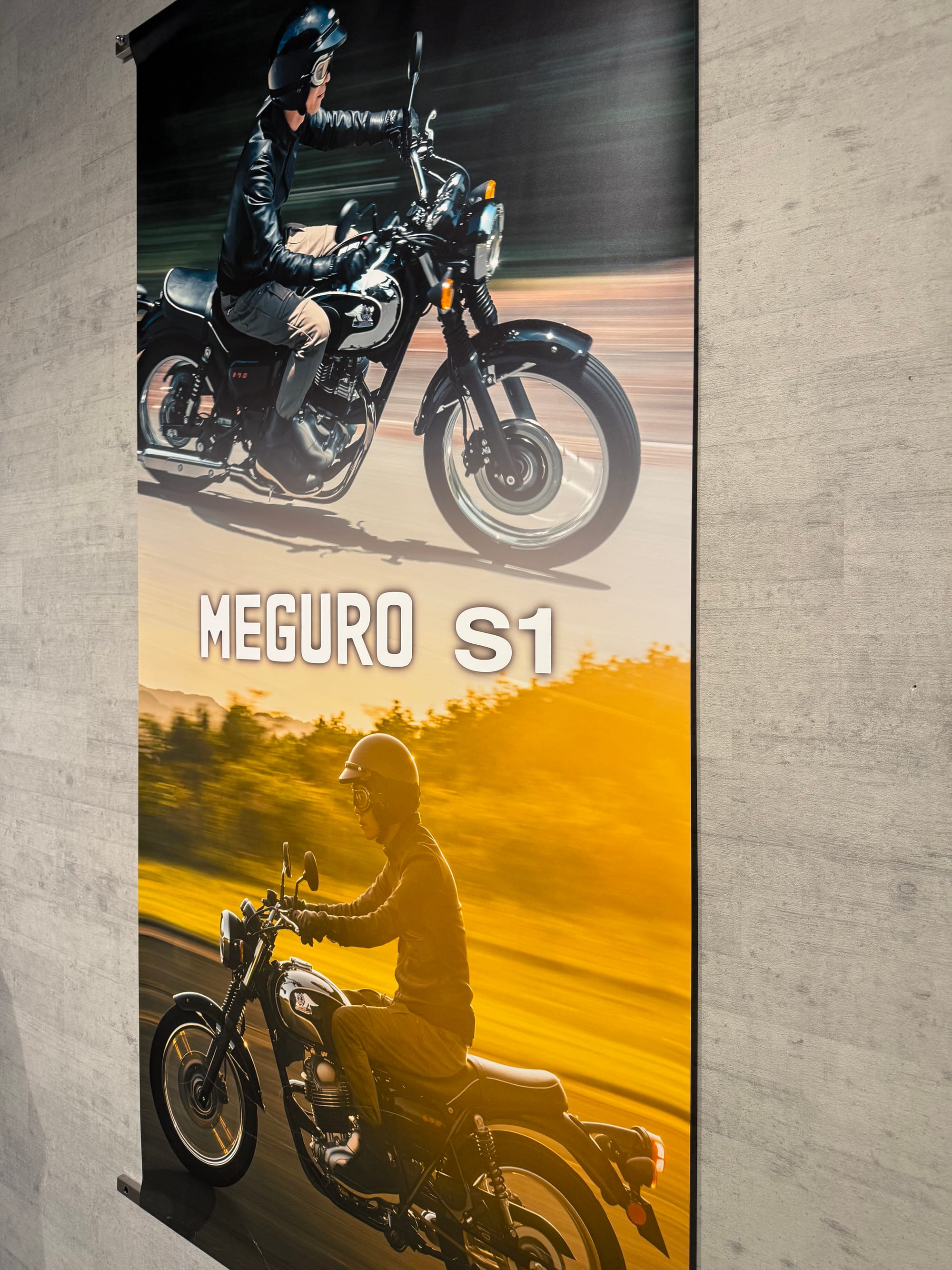 Promotional banner for the Kawasaki Meguro S1 motorcycle at Kawasaki Good Times World, featuring riders on the retro-styled bike against blurred road and countryside backdrops.