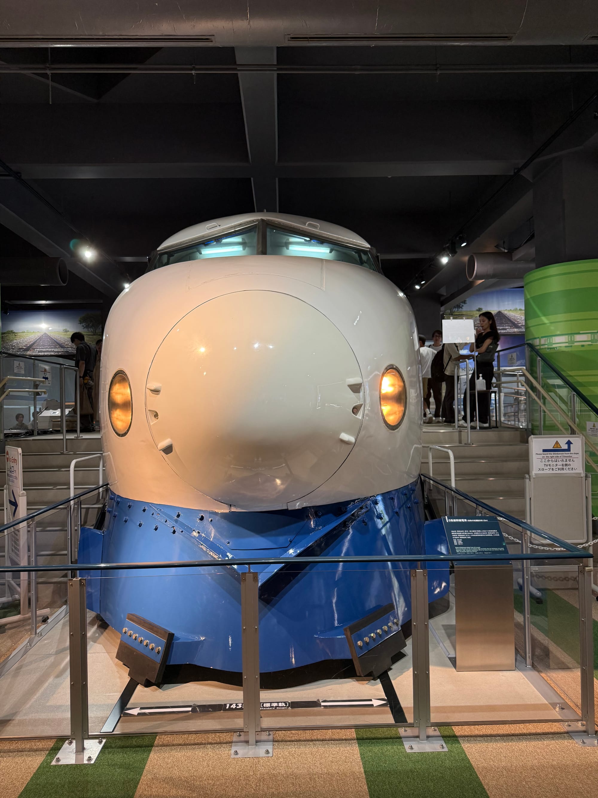 Front section of a 0 Series Shinkansen displayed at Kawasaki Good Times World in Kobe, showing the original bullet train's rounded nose, twin headlights and blue-and-white livery.