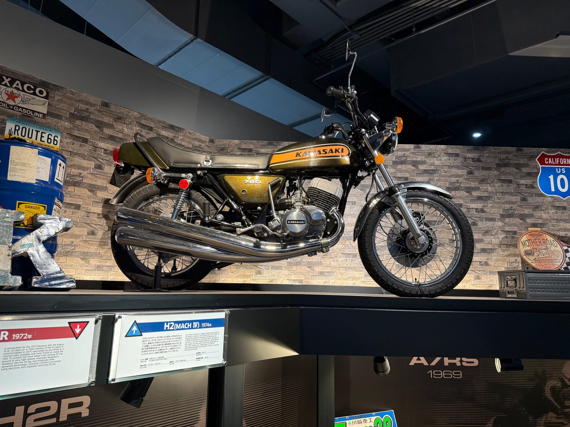 Kawasaki H2 Mach IV motorcycle displayed at Kawasaki Good Times World in Kobe, featuring a gold-and-black fuel tank, triple-cylinder two-stroke engine and distinctive triple exhausts.