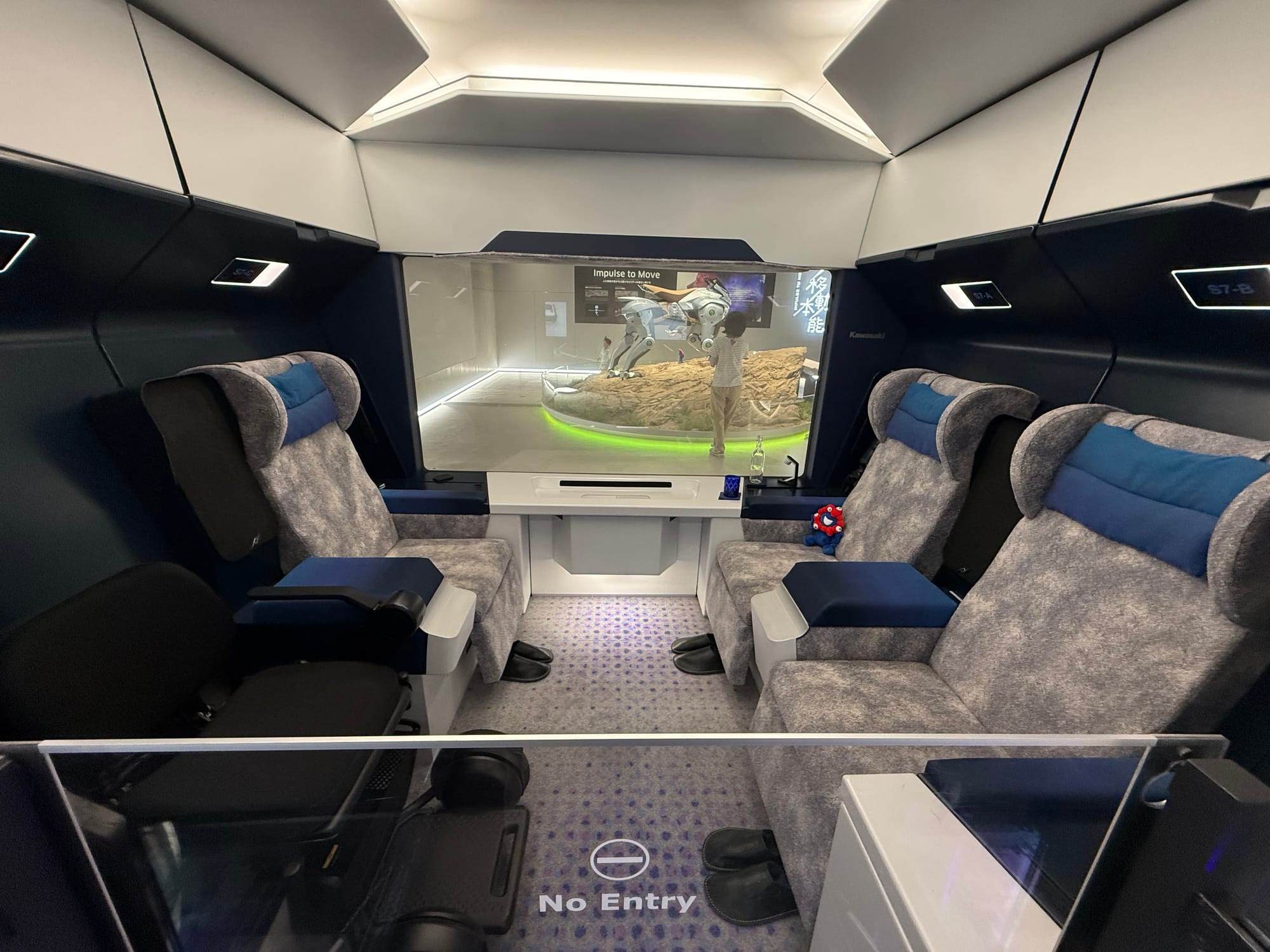 Future passenger cabin concept displayed at Kawasaki Good Times World, featuring spacious reclining seats, wheelchair access and a large panoramic window overlooking the exhibit hall.