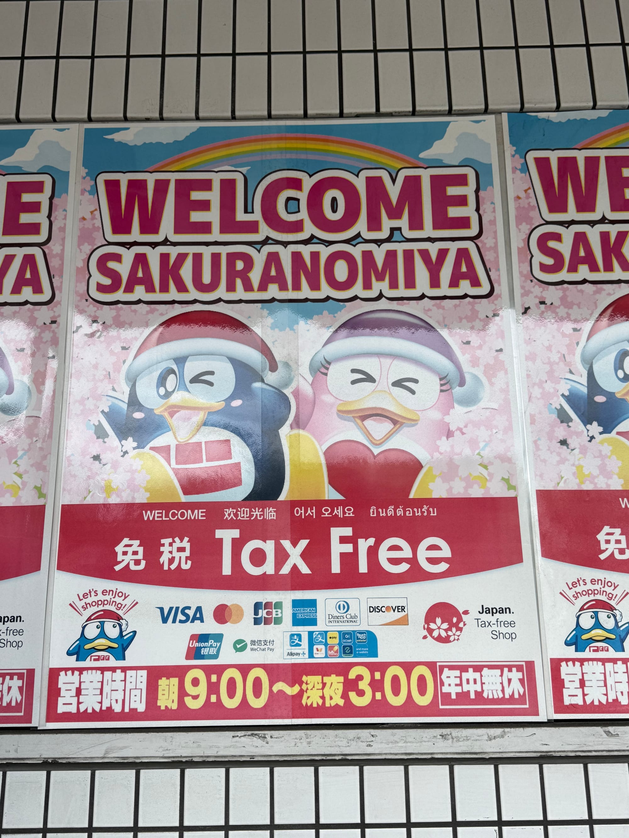 Welcome Sakuranomiya tax-free shopping sign in Osaka, featuring colourful Don Quijote penguin mascots, cherry blossom graphics, multilingual greetings and payment logos.