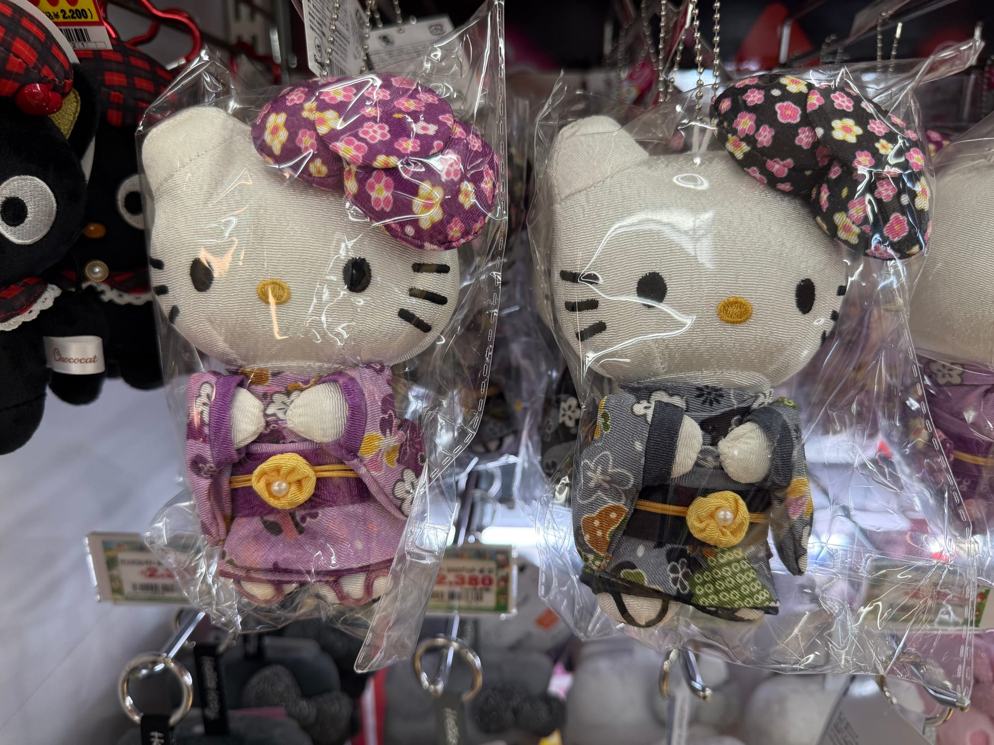 Hello Kitty plush keychains wearing traditional Japanese kimono are displayed for sale in a shop, featuring floral-patterned outfits in pink, purple, black and grey fabrics.