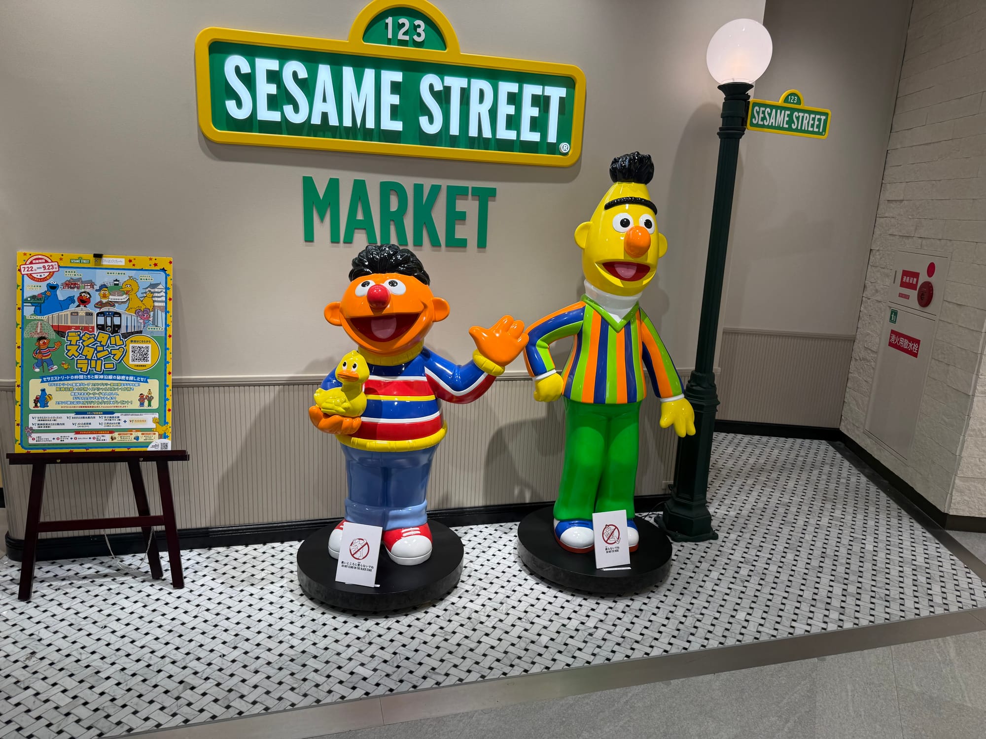 Ernie and Bert statues at Sesame Street Market in Osaka, standing beneath a Sesame Street sign and lamppost while posing beside a promotional display near the shop entrance.