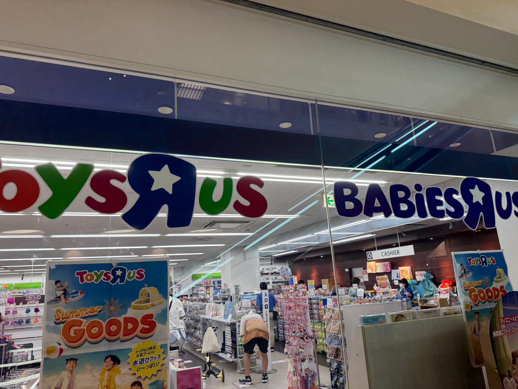 Toys “R” Us and Babies “R” Us storefront in Osaka, featuring the colourful logo above the entrance, with toy displays, shoppers and summer promotion signs visible inside.