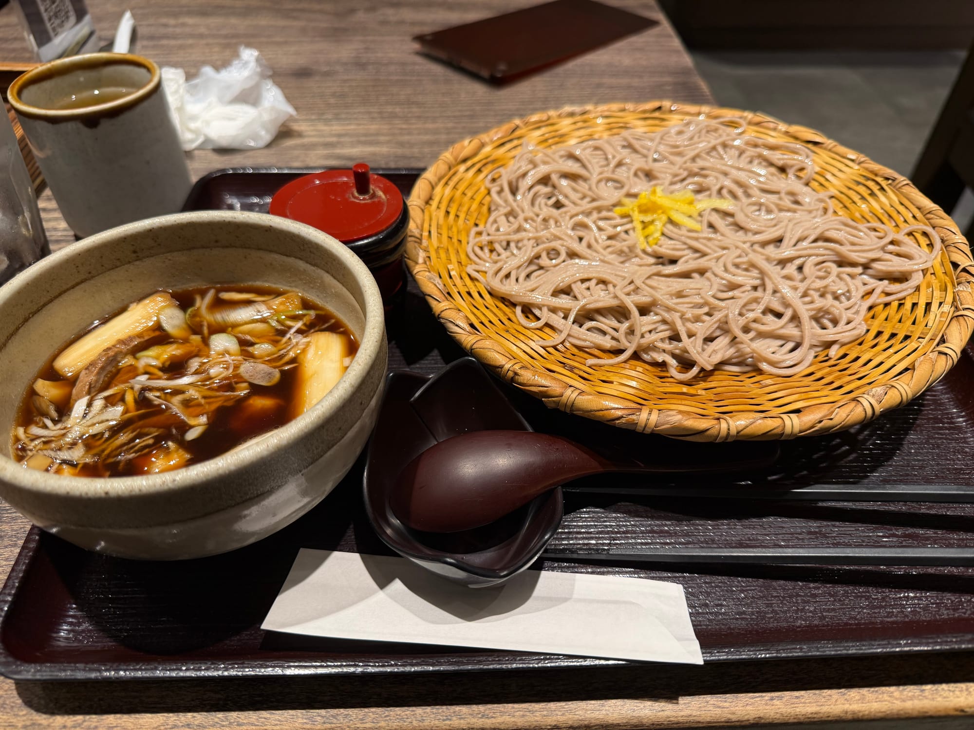 Basket of chilled soba noodles served with a hot dipping broth containing duck, mushrooms and leeks, presented on a lacquer tray with condiments and tea at a Japanese restaurant.