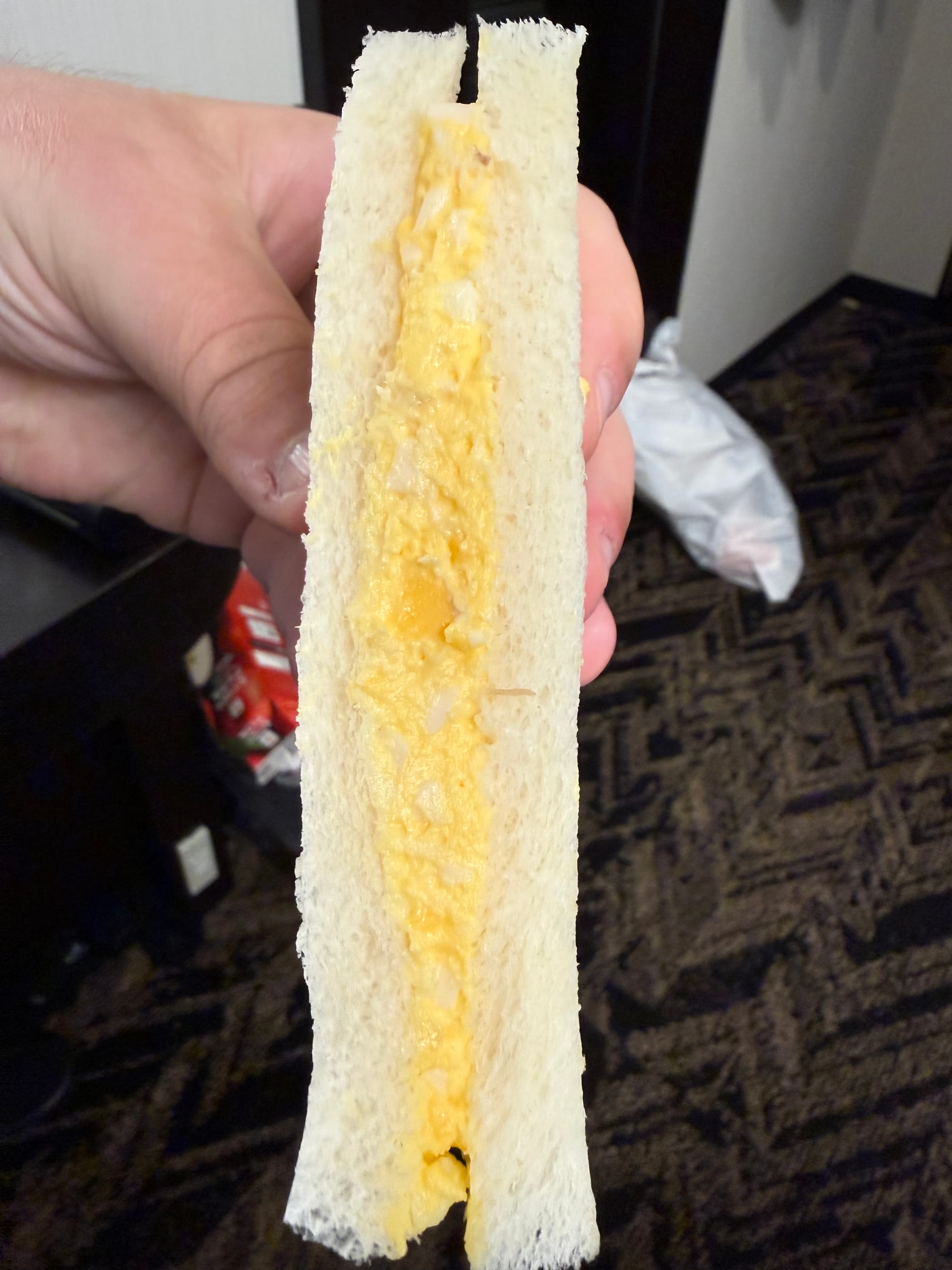 Japanese egg sandwich held by a visitor, featuring soft white bread filled with a creamy mixture of chopped boiled eggs and mayonnaise, photographed inside a hotel room.