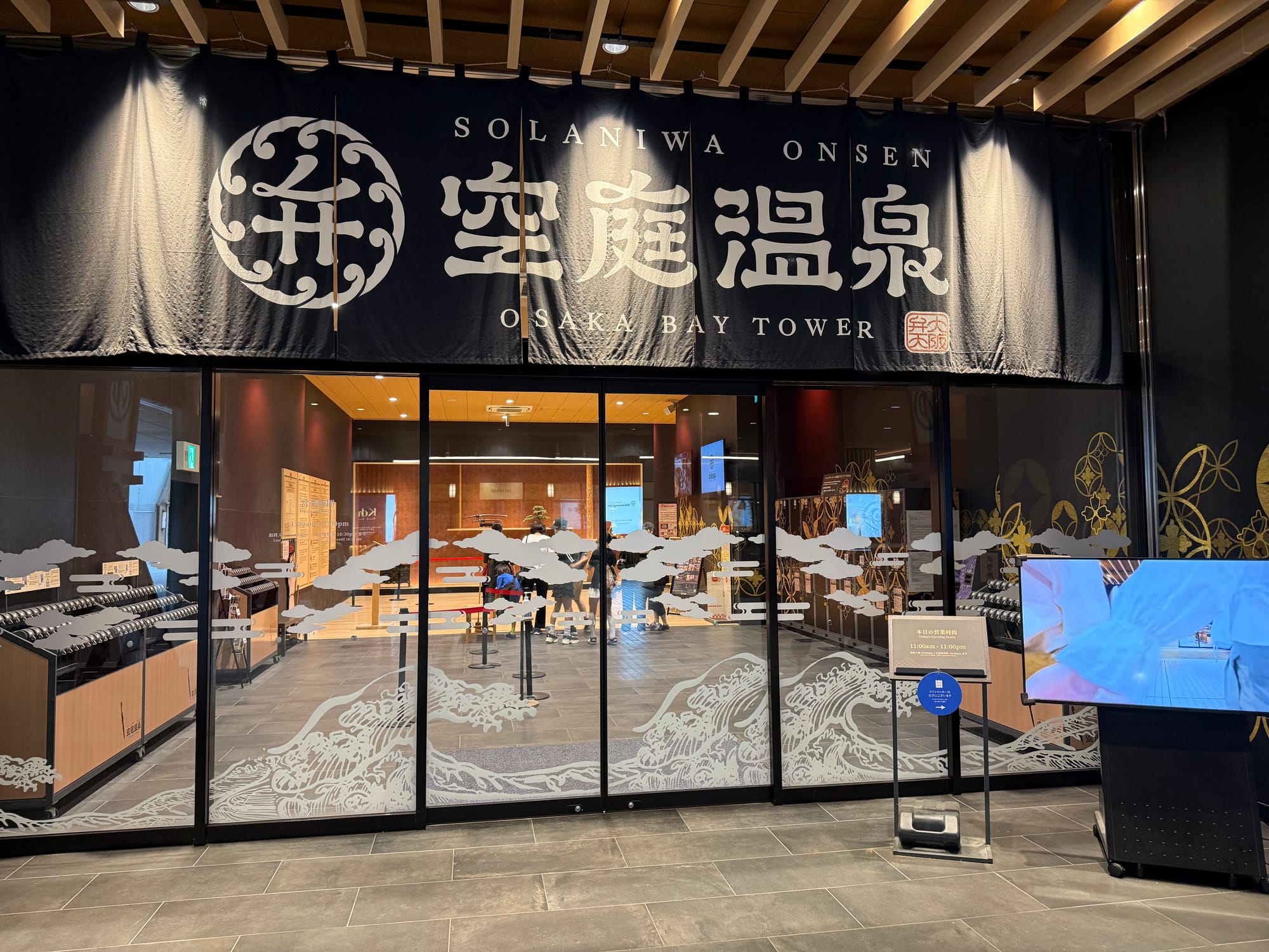 Entrance to Solaniwa Onsen Osaka Bay Tower, featuring a large traditional-style curtain with the facility name above glass doors decorated with Japanese wave and cloud motifs.