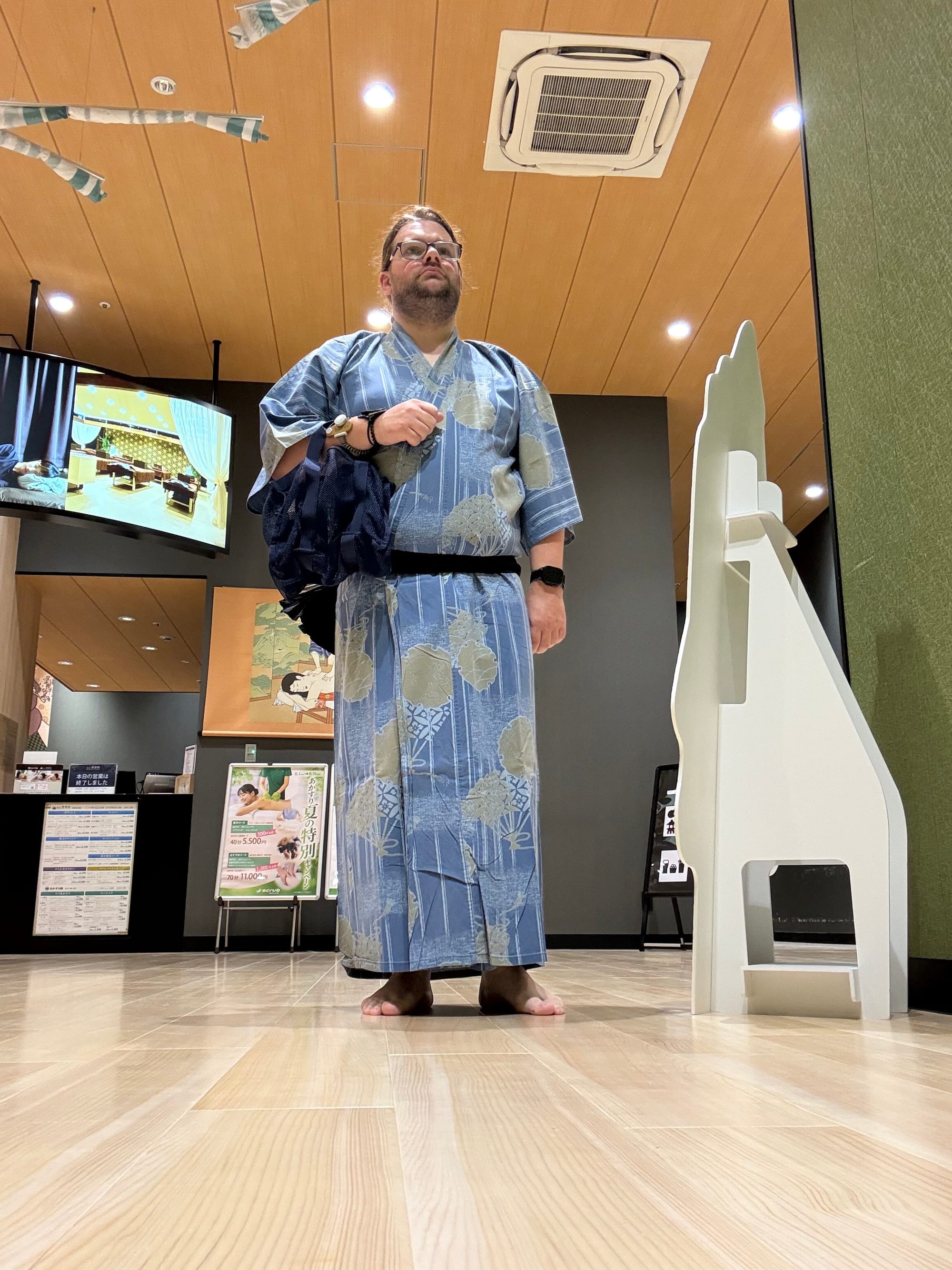 Visitor wearing a blue patterned yukata at Solaniwa Onsen Osaka Bay Tower, standing barefoot in the lobby area while carrying a bath bag provided by the facility.