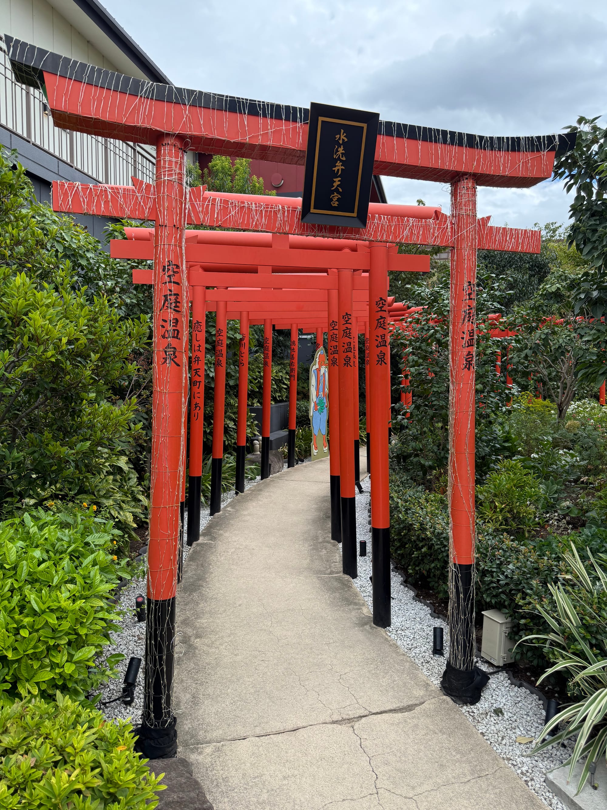 Pathway lined with vermilion torii gates in the rooftop garden at Solaniwa Onsen Osaka Bay Tower, winding through landscaped greenery beneath an overcast sky.