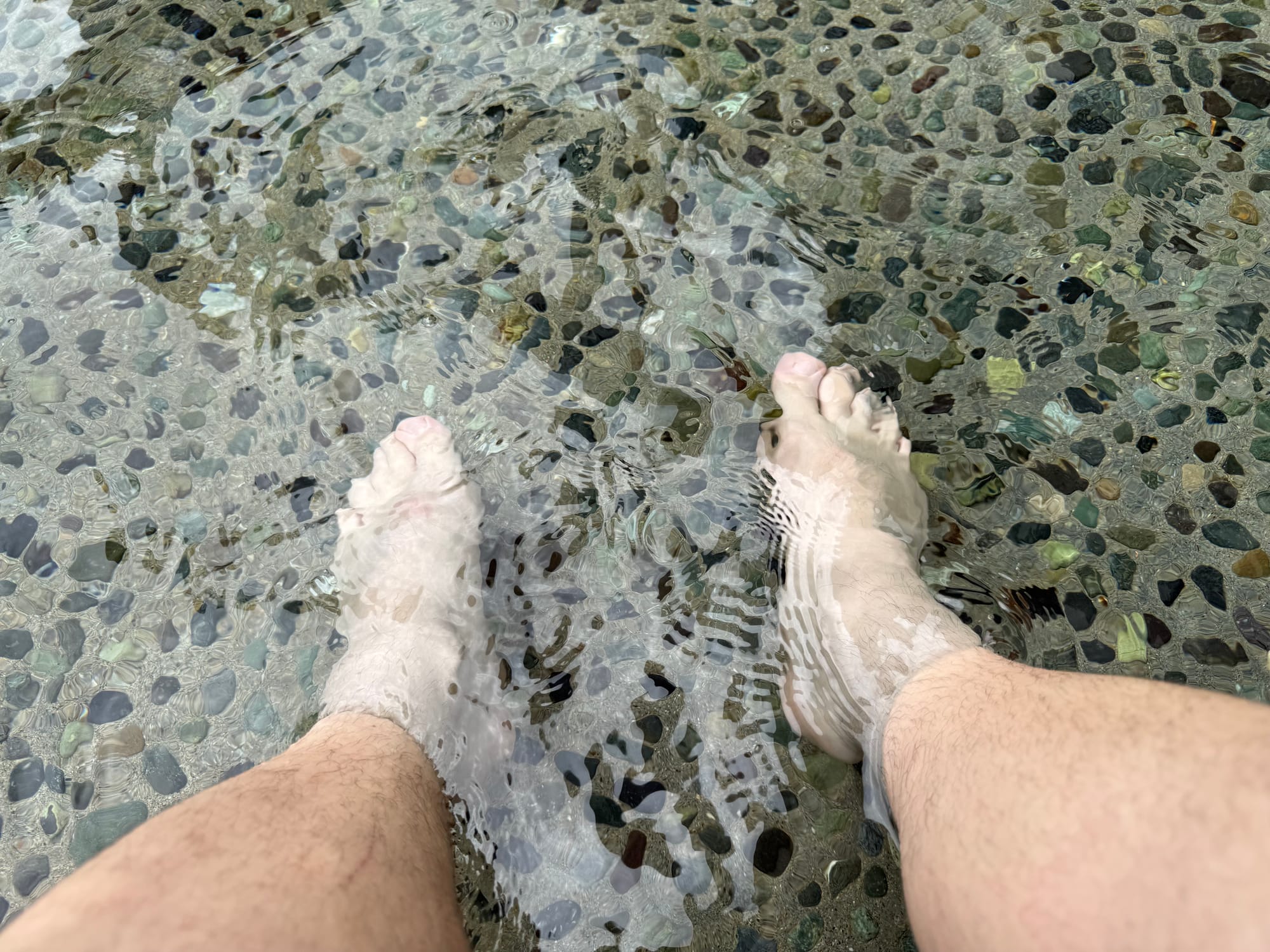 Feet submerged in the cooling foot spa at Solaniwa Onsen Osaka Bay Tower, with clear water flowing over a pebble-lined basin designed for relaxation between hot baths.