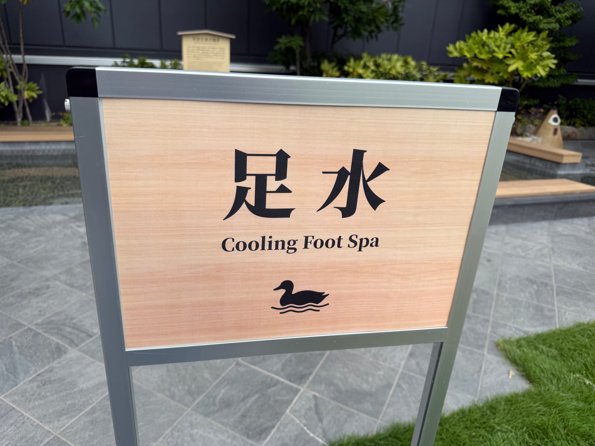 Sign for a cooling foot spa in the rooftop garden at Solaniwa Onsen Osaka Bay Tower, with the shallow footbath, landscaped greenery and relaxation area visible behind.