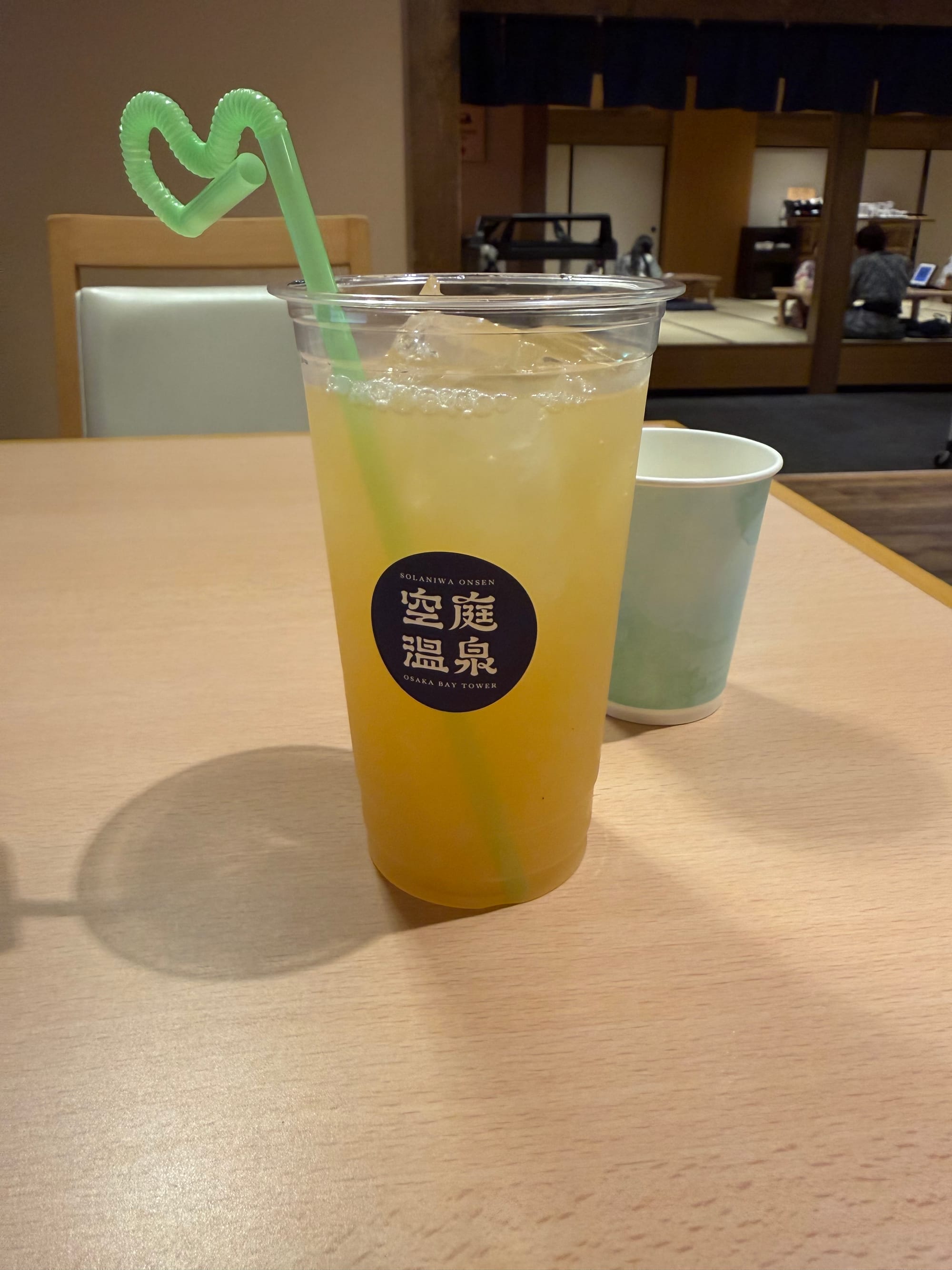 Cup of chilled apple juice served at Solaniwa Onsen Osaka Bay Tower, with ice cubes and a heart-shaped green straw, placed on a table in a relaxation area.