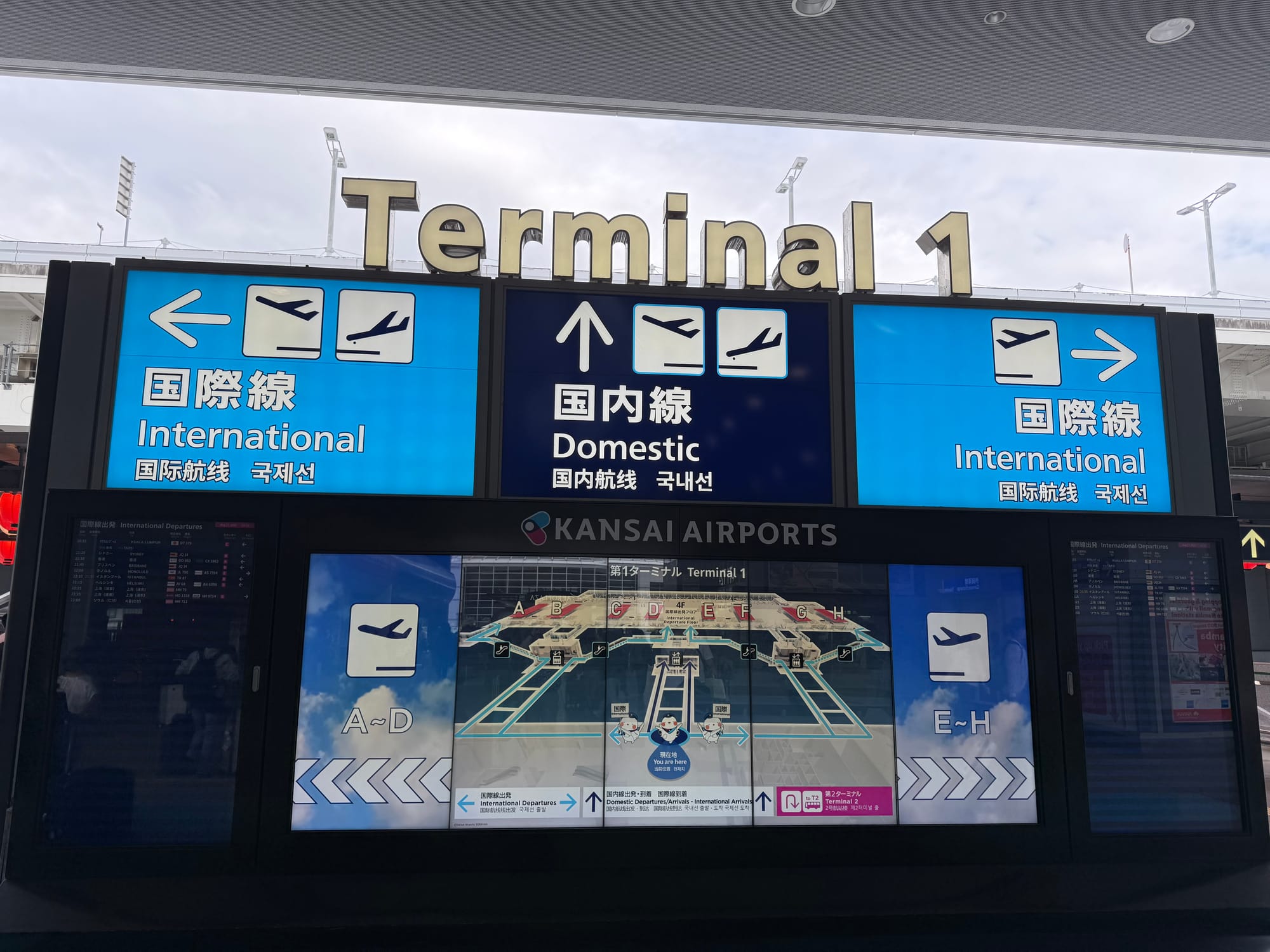 Terminal 1 directional sign at Kansai International Airport, showing routes to domestic and international departures, with terminal maps and flight information displays below.