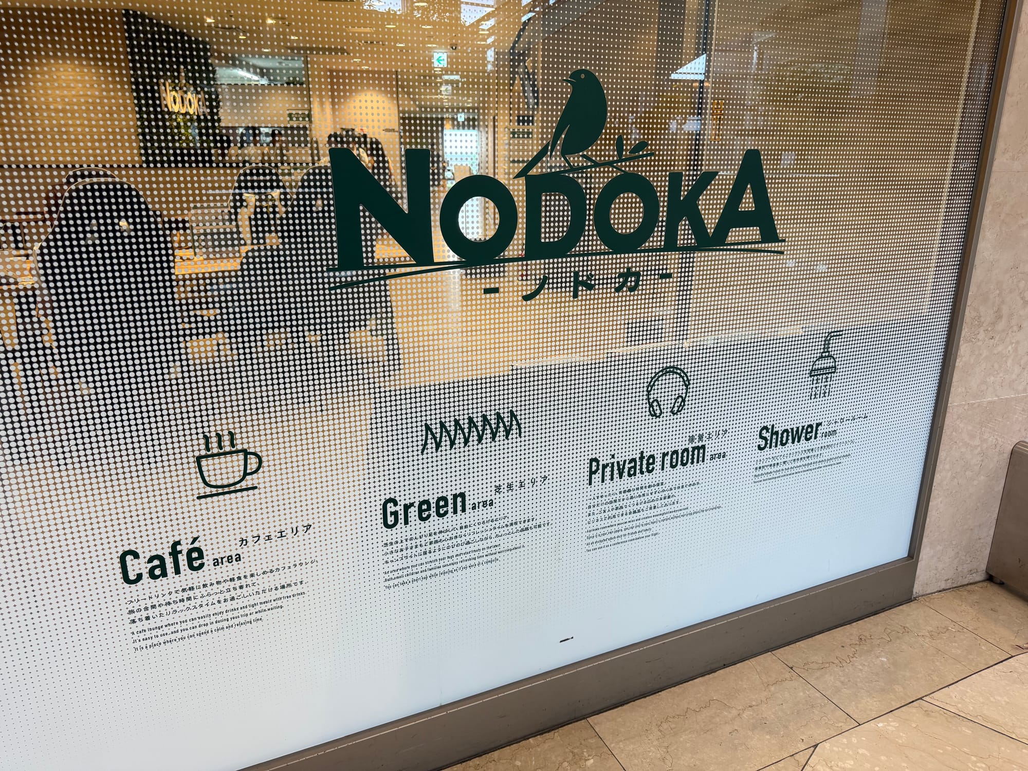 Entrance sign for the Nodoka airport lounge at Kansai International Airport, showing facilities including a café, green area, private rooms and shower rooms behind a glass wall.