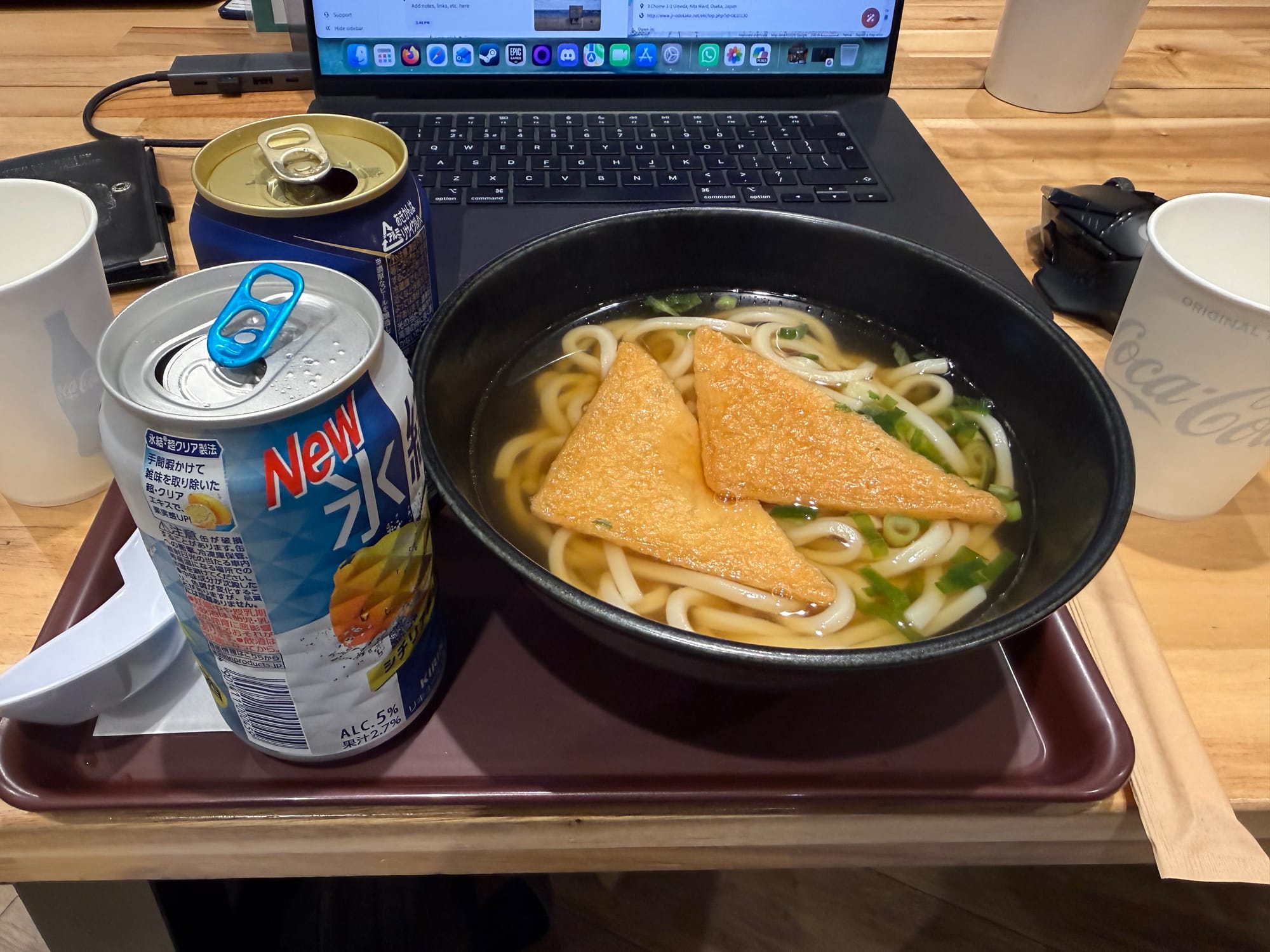 Bowl of kitsune udon topped with fried tofu served in the Nodoka lounge at Kansai International Airport, alongside canned drinks, chopsticks and a laptop on a wooden table.