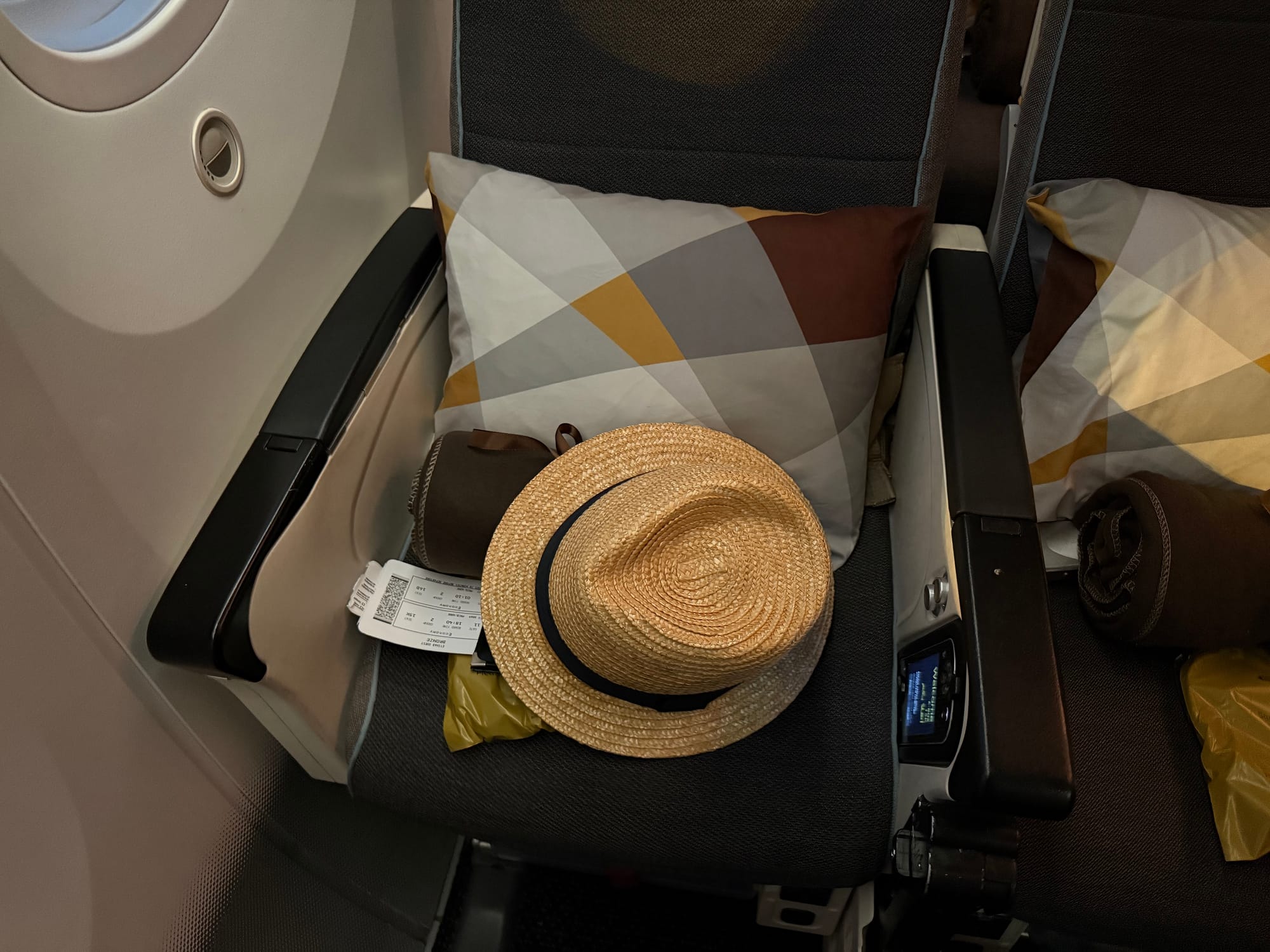Straw hat resting on a Premium Economy seat aboard an Etihad Airways Boeing 787-9, partially covering a boarding pass, with a pillow and blanket arranged for the flight.