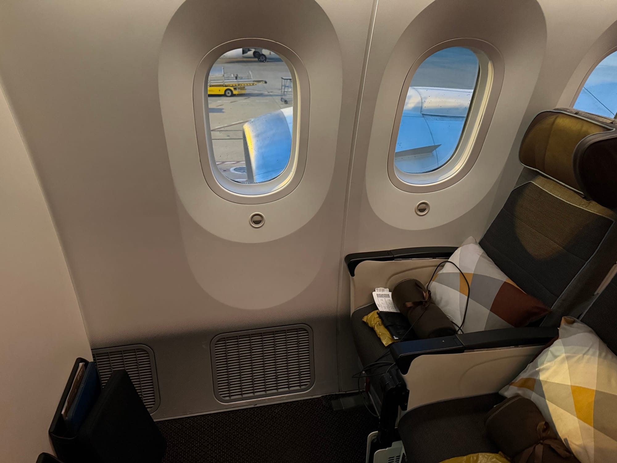 Premium Economy seat aboard an Etihad Airways aircraft, showing the extra legroom row beside two windows, with the engine and airport apron visible outside.