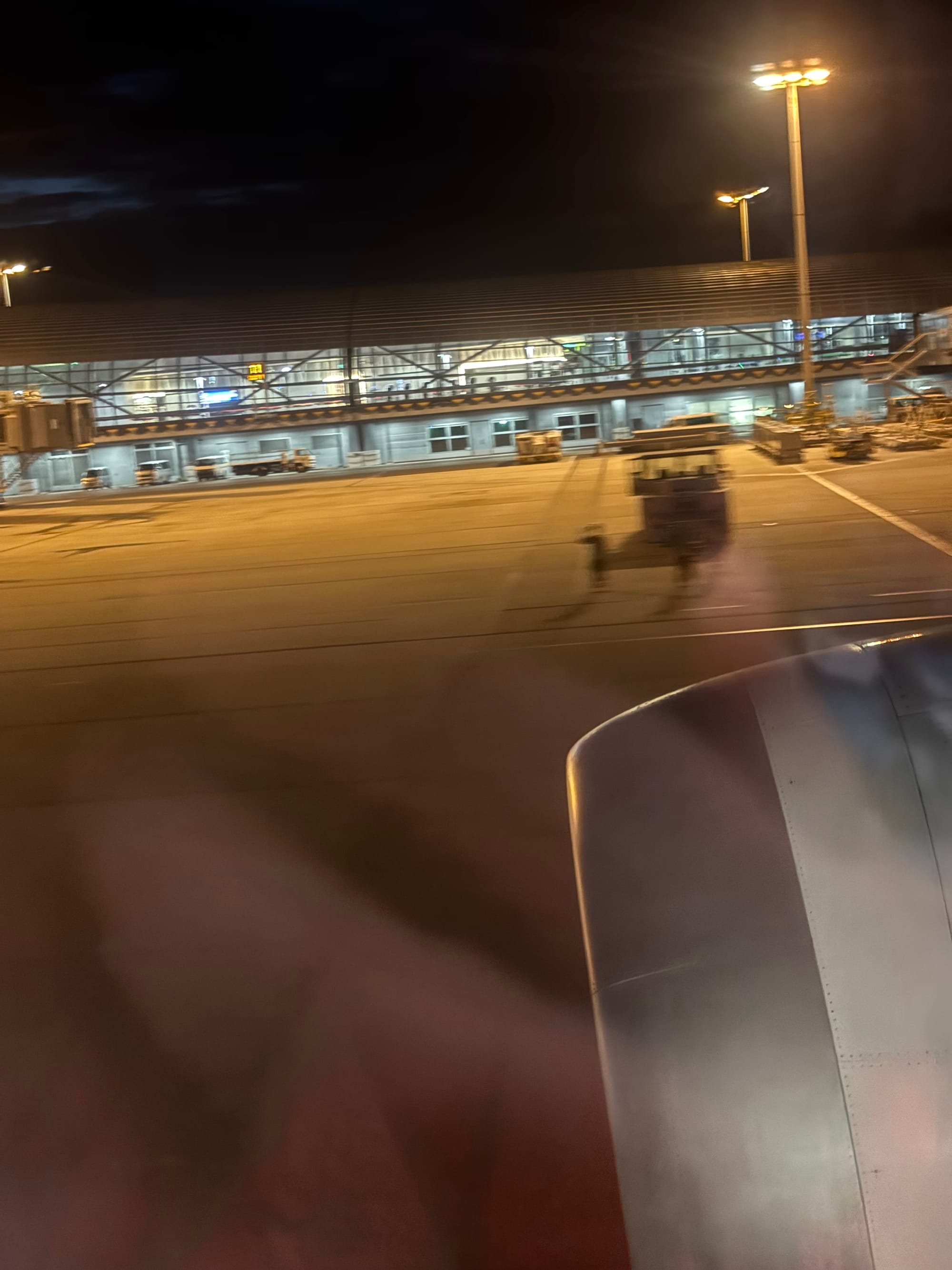 Ground crew wave from beside a pushback tug at Kansai International Airport at night, viewed from an Etihad Airways aircraft window with the engine and terminal visible.