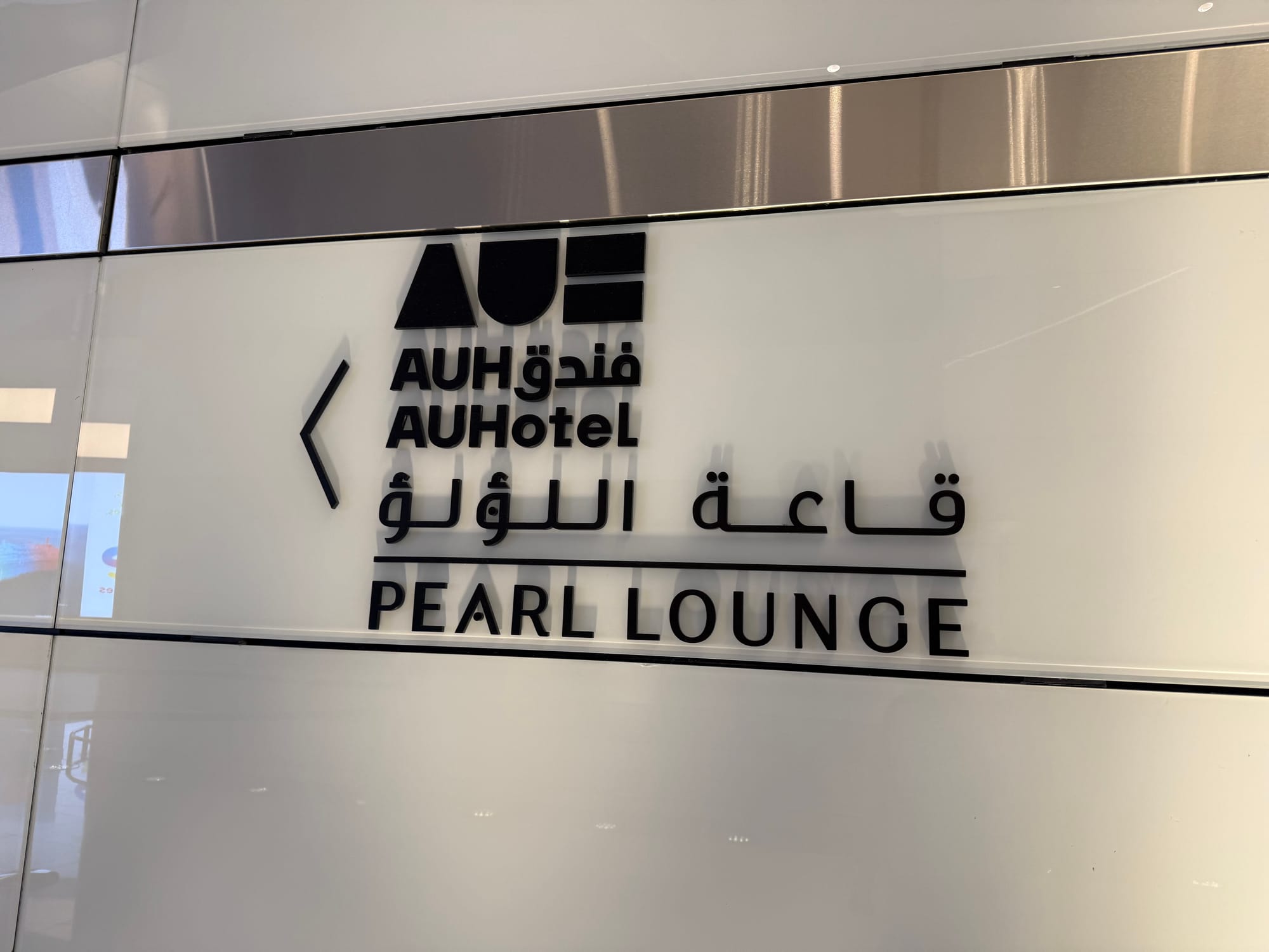 Sign for the Pearl Lounge at Abu Dhabi International Airport, displaying directions to the lounge beside the AUHotel branding on a white wall with Arabic and English text.