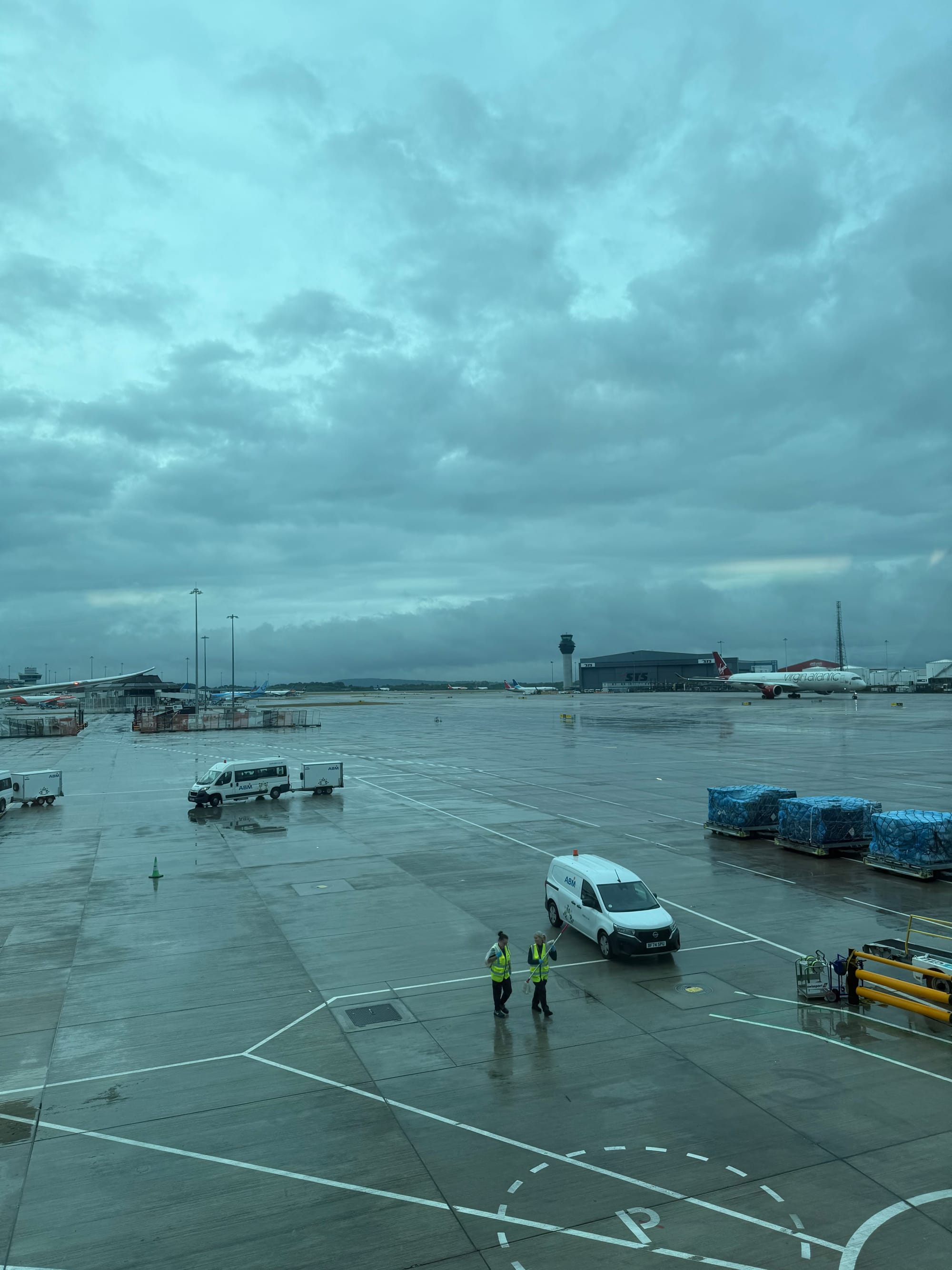 Rain-soaked apron at Manchester Airport viewed through terminal windows, with ground vehicles, aircraft stands, the control tower and low clouds stretching across the sky.