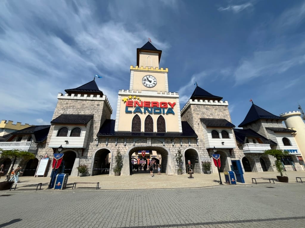 Entrance to Energylandia theme park in Poland, featuring a castle-style façade with towers, a clock tower and the park's colourful sign beneath a bright sky.