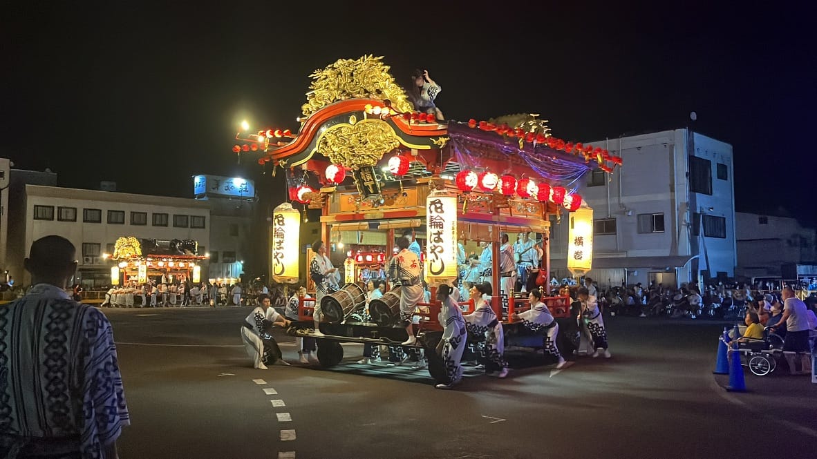 Illuminated festival float decorated with lanterns and carvings, paraded through the streets of Kazuno at night, accompanied by musicians and participants in traditional dress.
